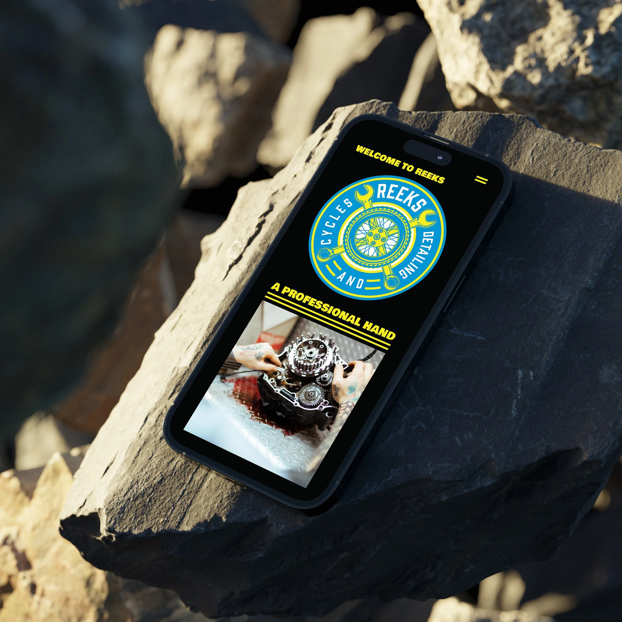 Smartphone displaying a website for 'Reeks Cycles and Detailing' with a logo and image of hands working on a mechanical part, placed on a rocky surface.