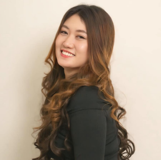 A smiling young woman with long, wavy brown hair, wearing a black top, standing against a plain wall.