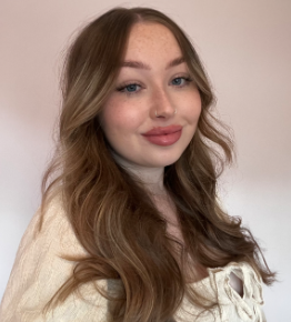 A young woman with long wavy light brown hair, wearing a cream-colored sweater, smiling softly at the camera against a plain background.