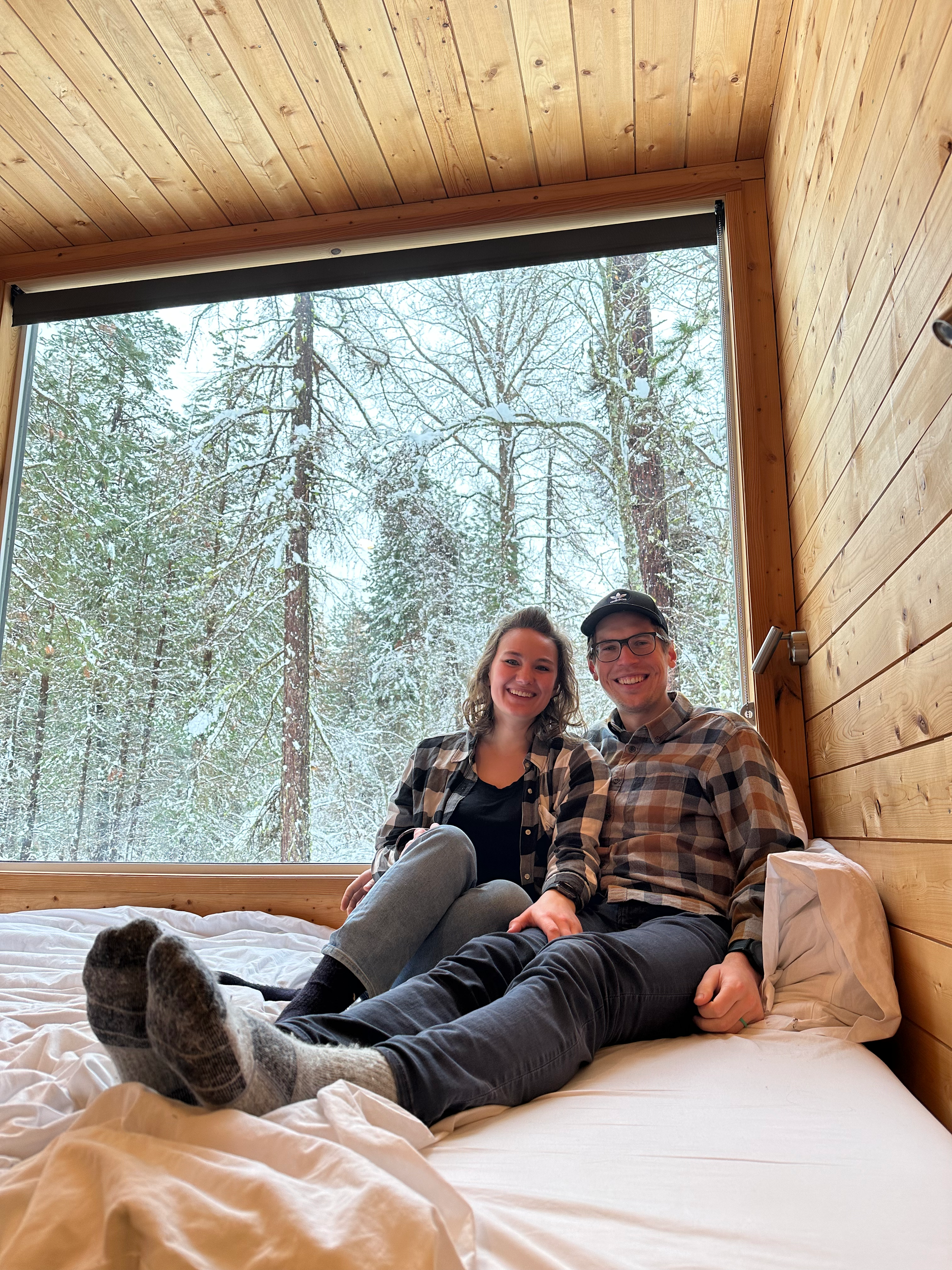 Three people sitting and lying on a bed inside a wooden cabin with a large window, overlooking snow-covered trees outside.