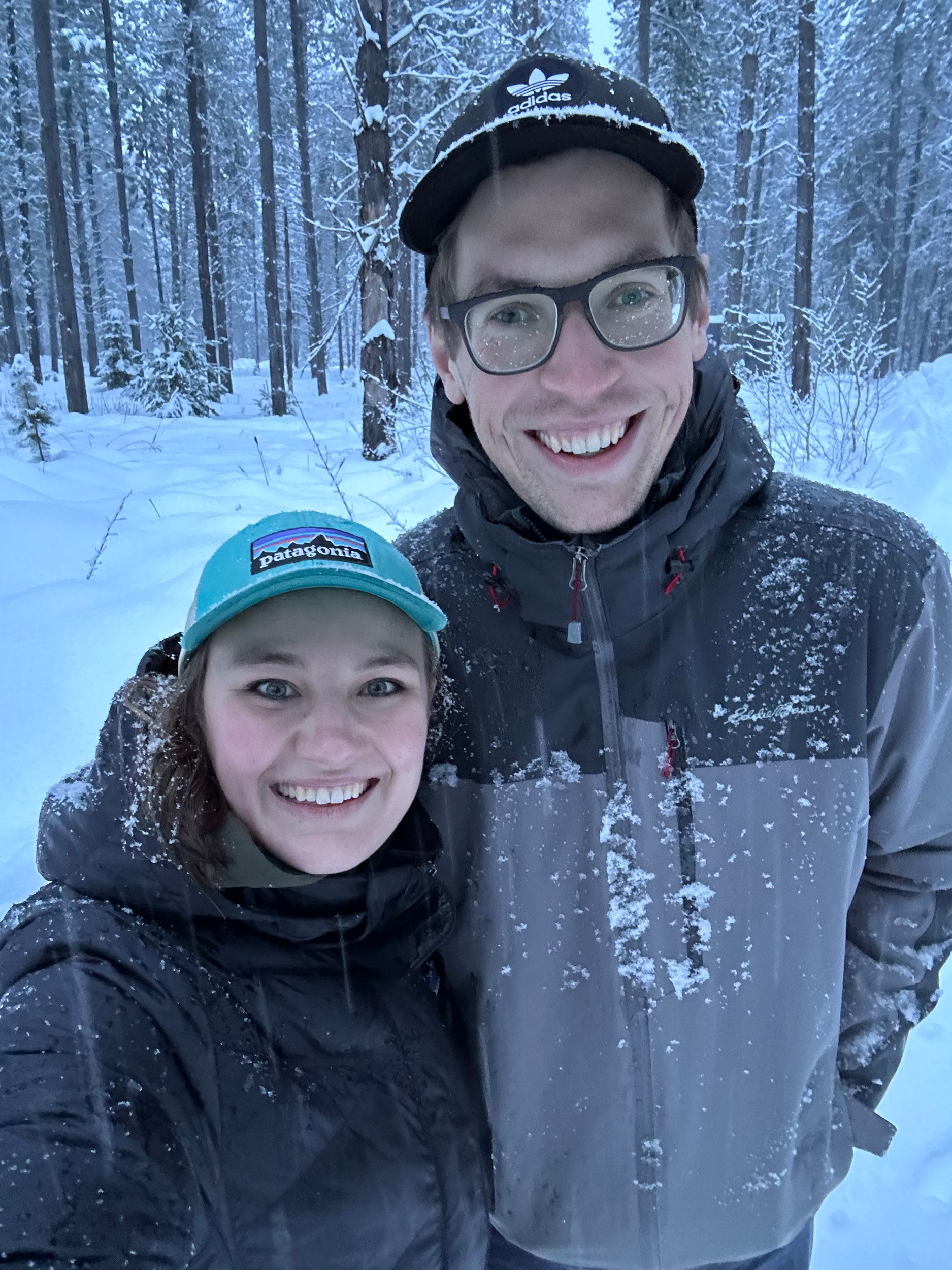 Two smiling people in winter clothing standing in snowy forest, taking a selfie together.
