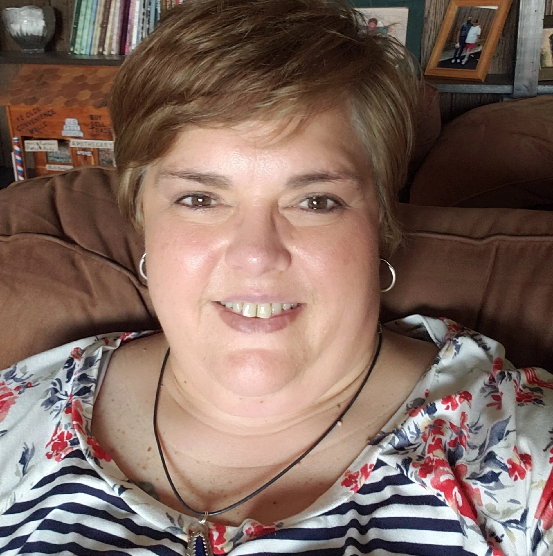 A woman with short brown hair, wearing a white top with red and blue floral and striped patterns, is smiling and seated on a brown leather couch in a room with bookshelves and family photos in the background.