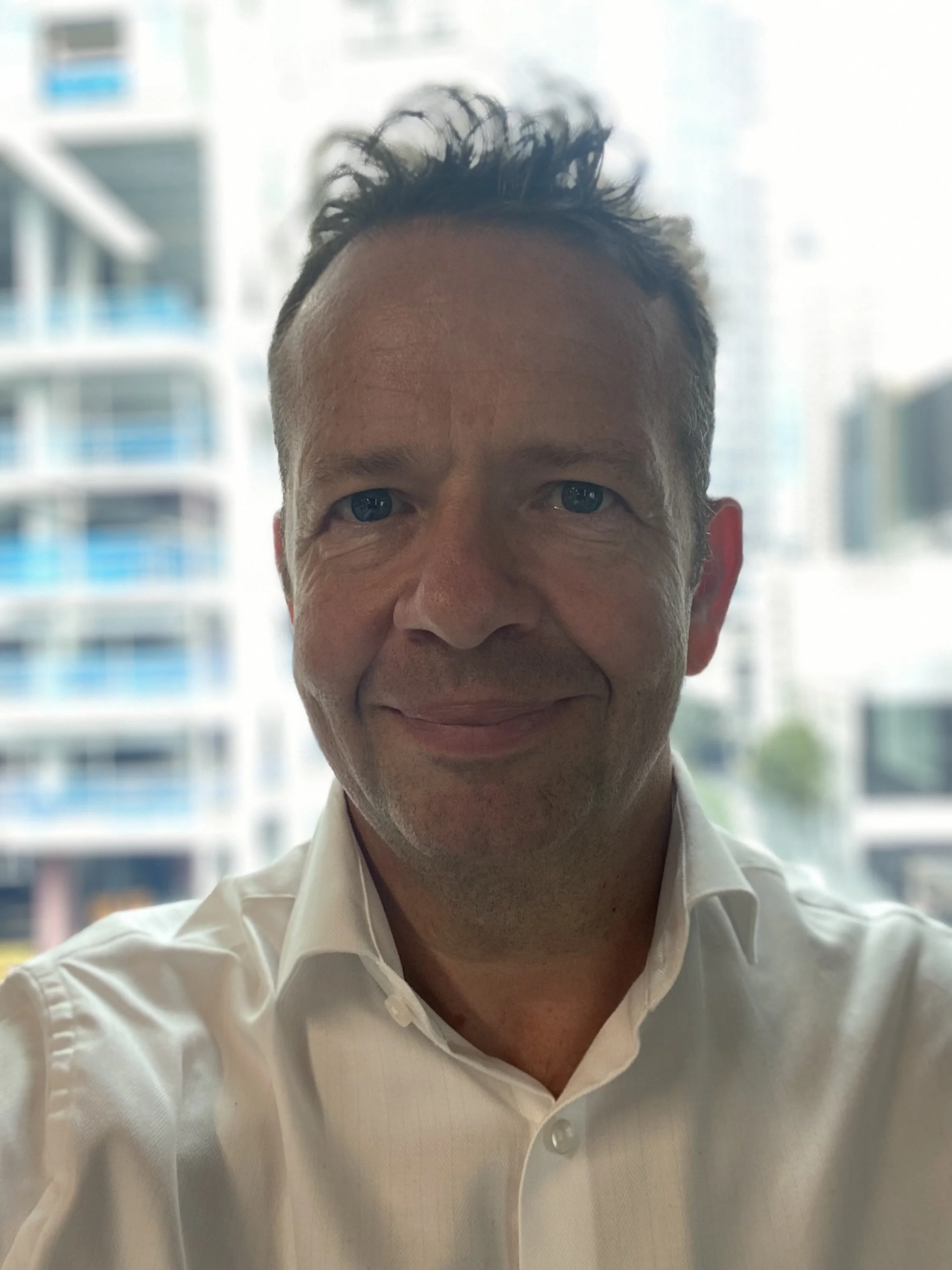 A man taking a selfie indoors with a cityscape of tall buildings with glass windows in the background.