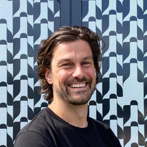 Hamish Howard headshot showing his brown hair and beard standing in front of patterned blue and black background.