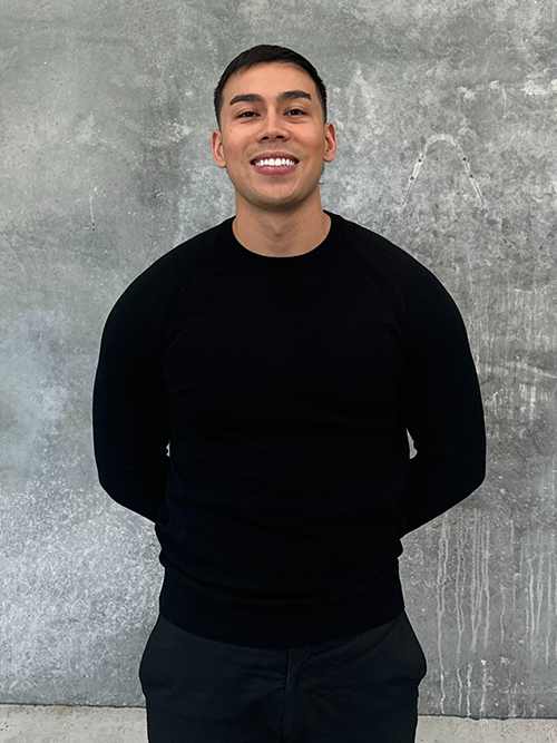 Smiling man in black sweater standing against a gray textured wall.