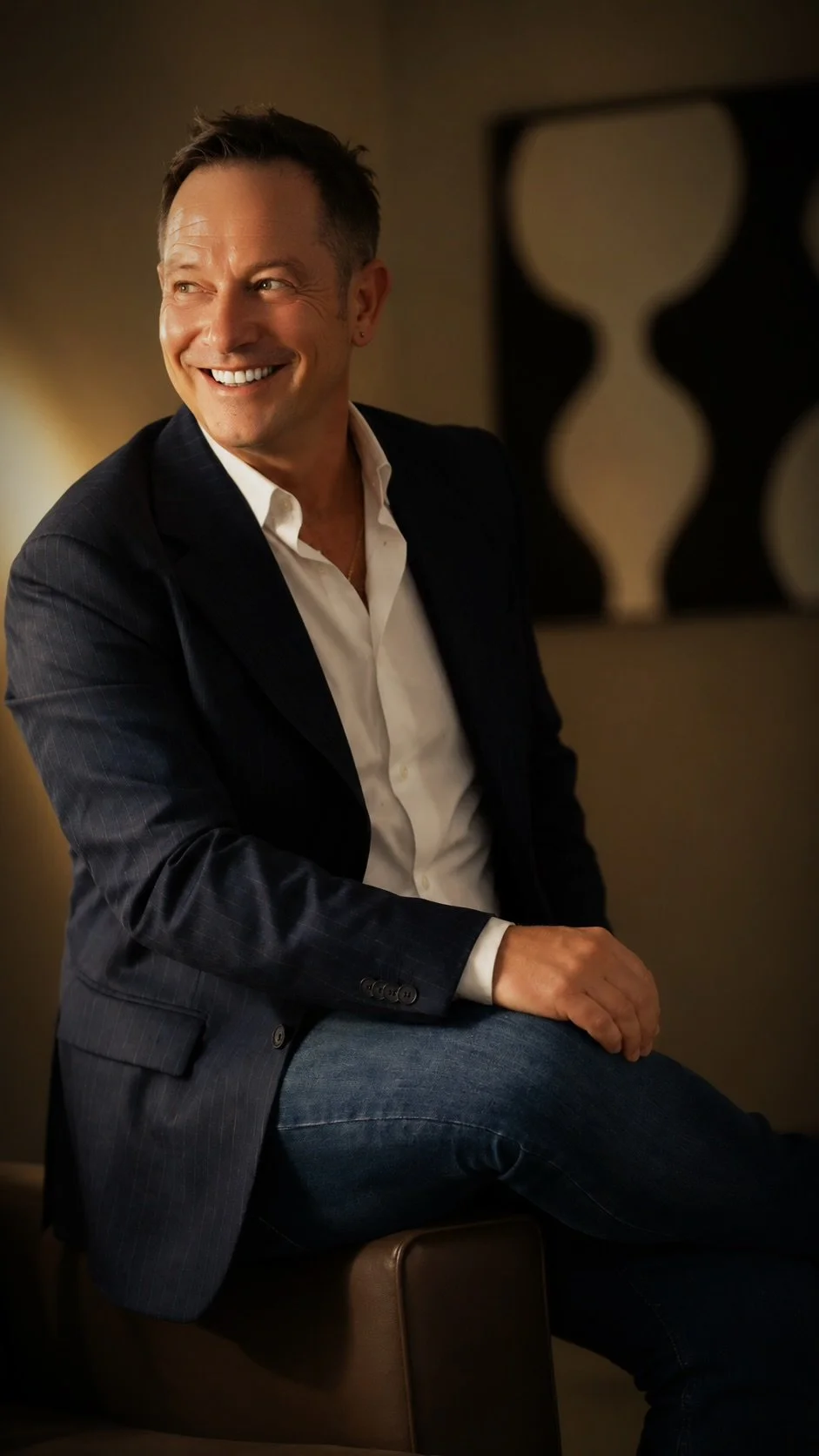 Hombre sonriente usando traje negro y camisa blanca, sentado en un sillón en un ambiente interior.