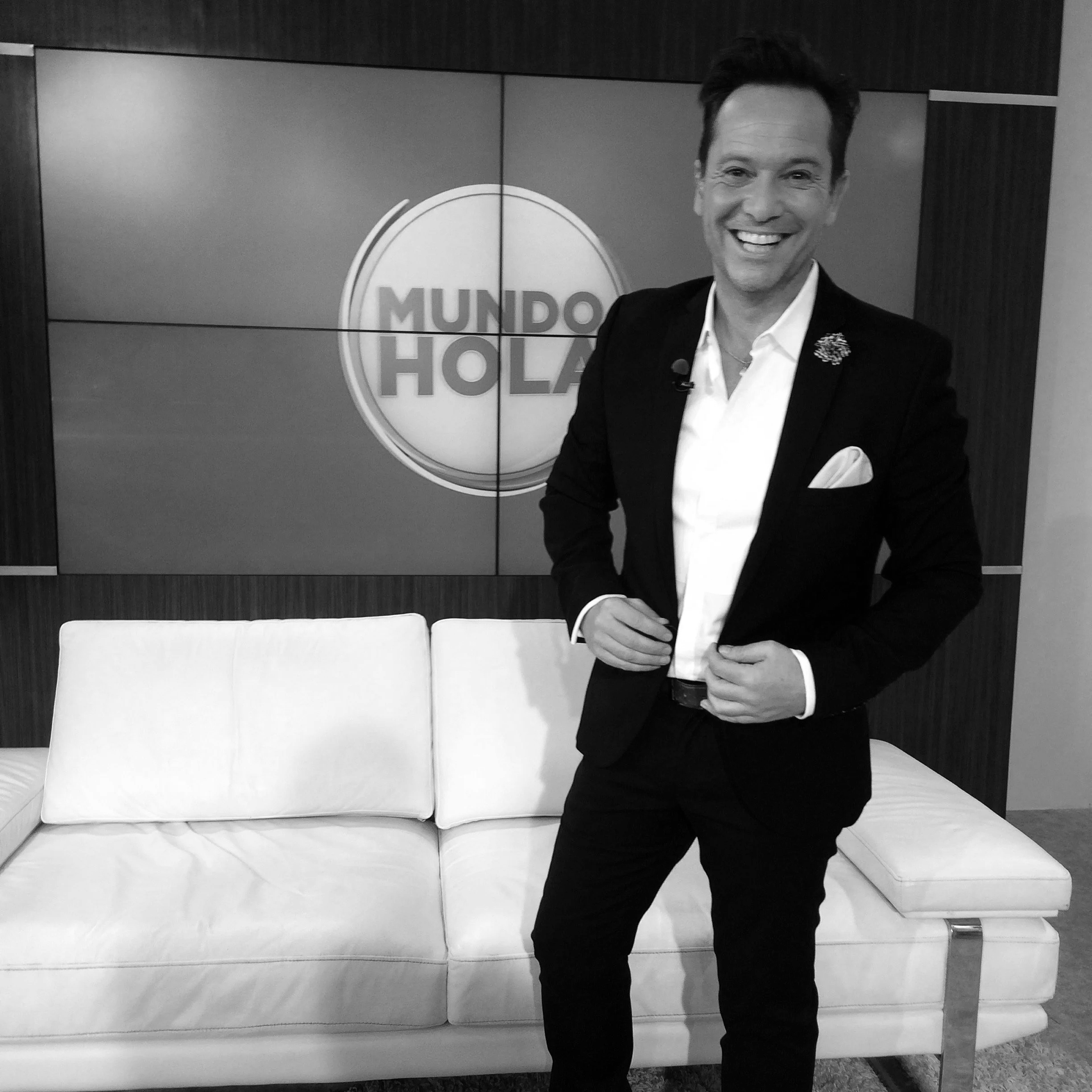 Hombre con traje negro y camisa blanca sonriendo, posando frente a un fondo de paredes oscuras y un sofá blanco.