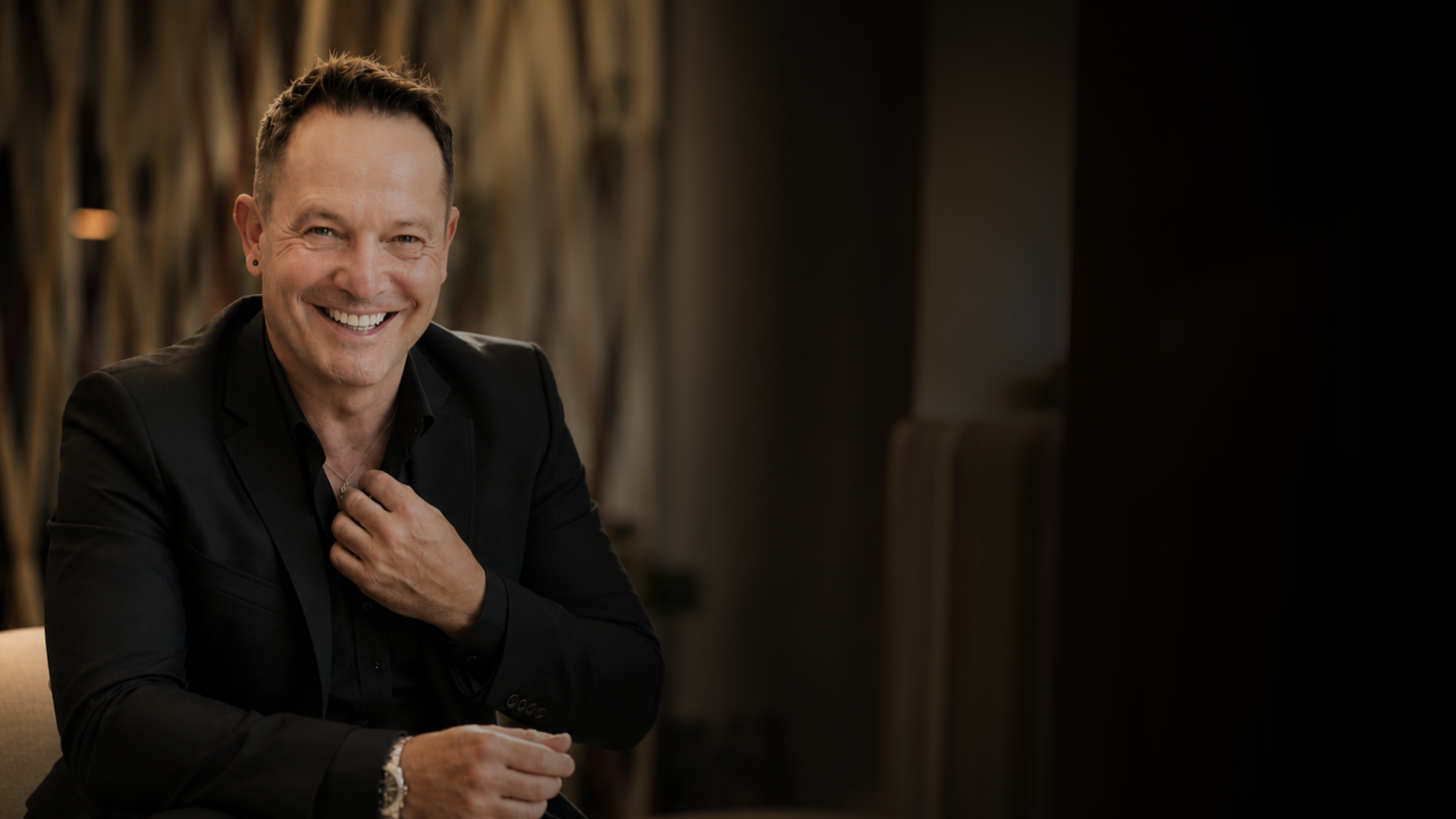 Hombre sonriendo, vestido con saco negro, en un ambiente con decoración moderna y suave iluminación.