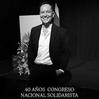 Hombre sonriendo en un escenario con un fondo negro, vestido con traje y camisa blanca, frente a un cartel que dice '40 años congreso nacional solidario' y decoraciones florales.