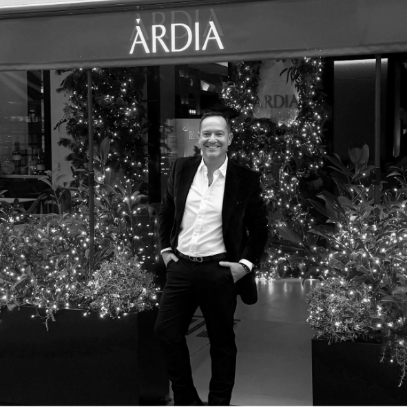 Hombre con traje negro y camisa blanca posando frente a una tienda llamada 'ÁRDIA', decorada con árboles de Navidad y luces.