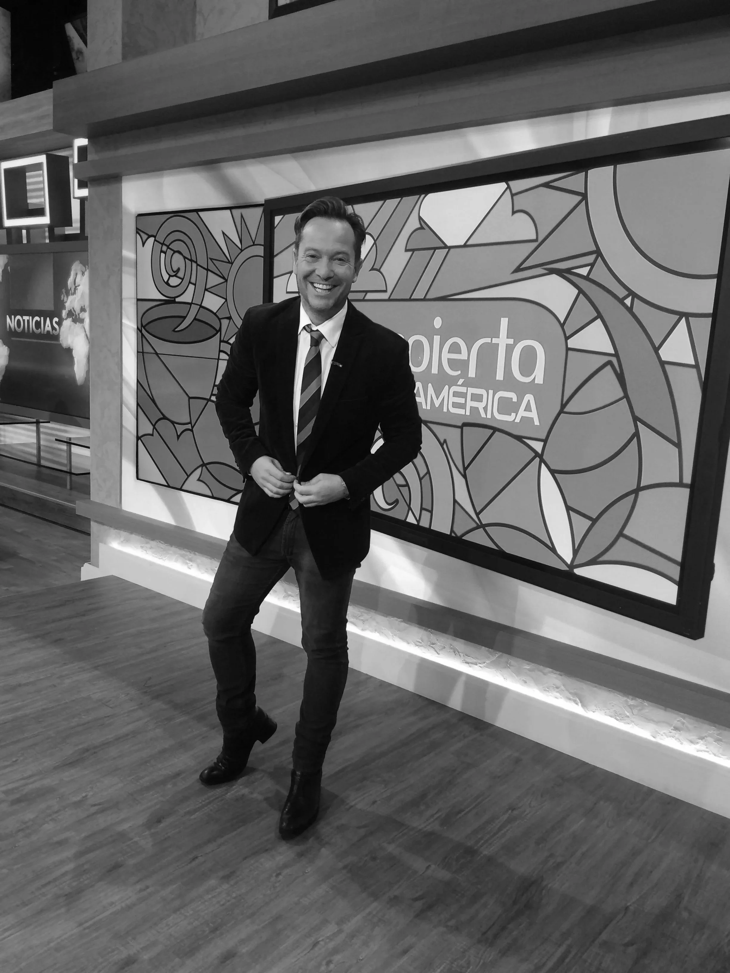 Hombre de traje y corbata sonriendo en un estudio de noticias con un cartel que dice 'abierto América' y un logotipo de un sol con nubes en el fondo.