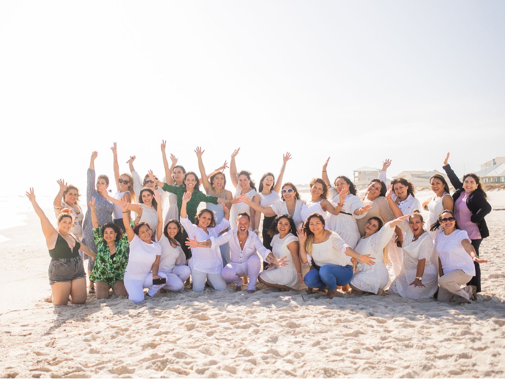 Grupo de mujeres y un hombre sonrientes y con los brazos abiertos en la playa, celebrando con fondo de arena y casas en la costa.