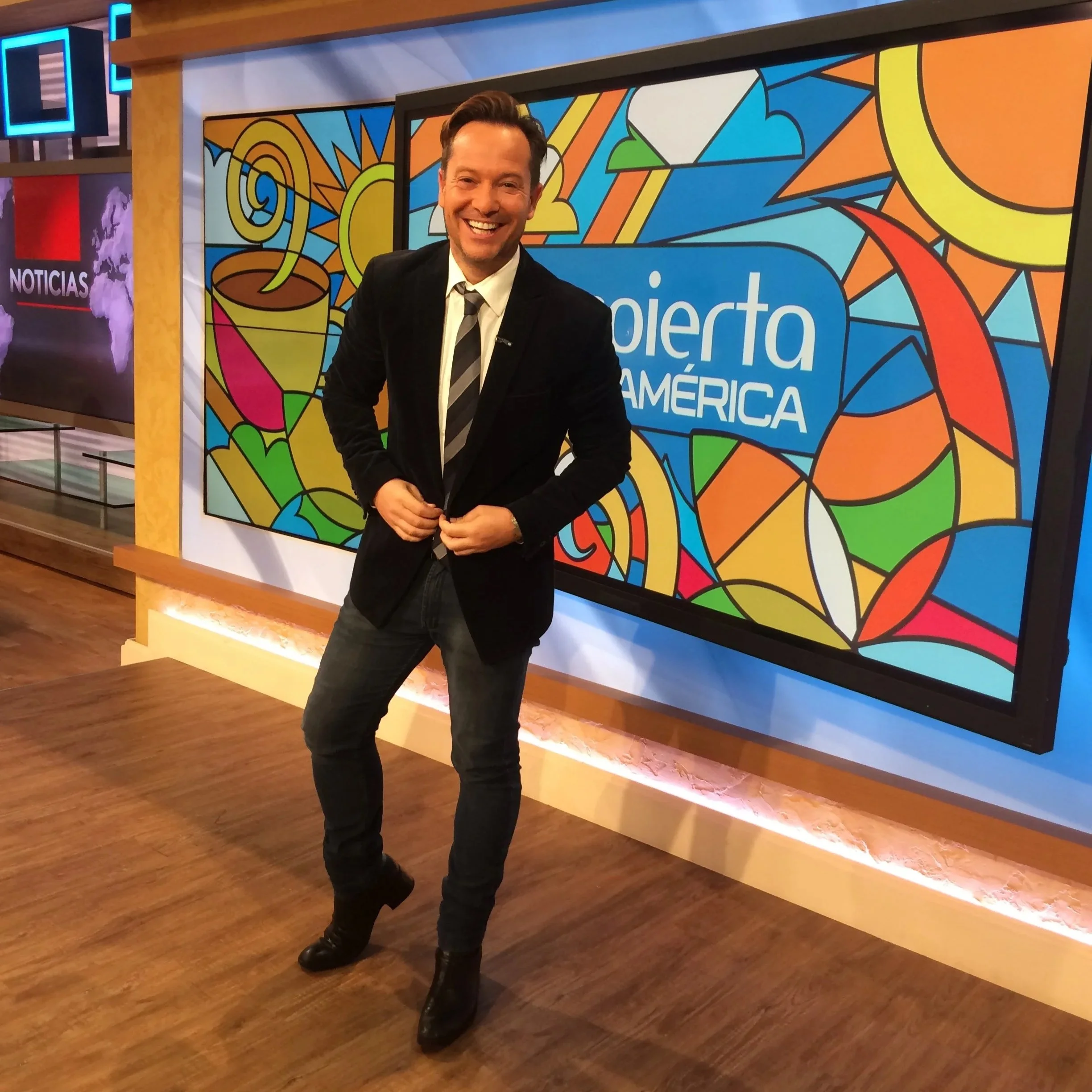 Hombre sonriendo con traje oscuro y camisa blanca, en fondo con pantallas y un mural colorido que dice 'Radio Ronda América'.