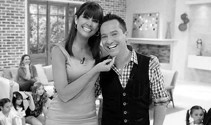 Dos niños, una niña y un niño, sonriendo en un escenario de programa de televisión, con otros niños y adultos en el fondo.