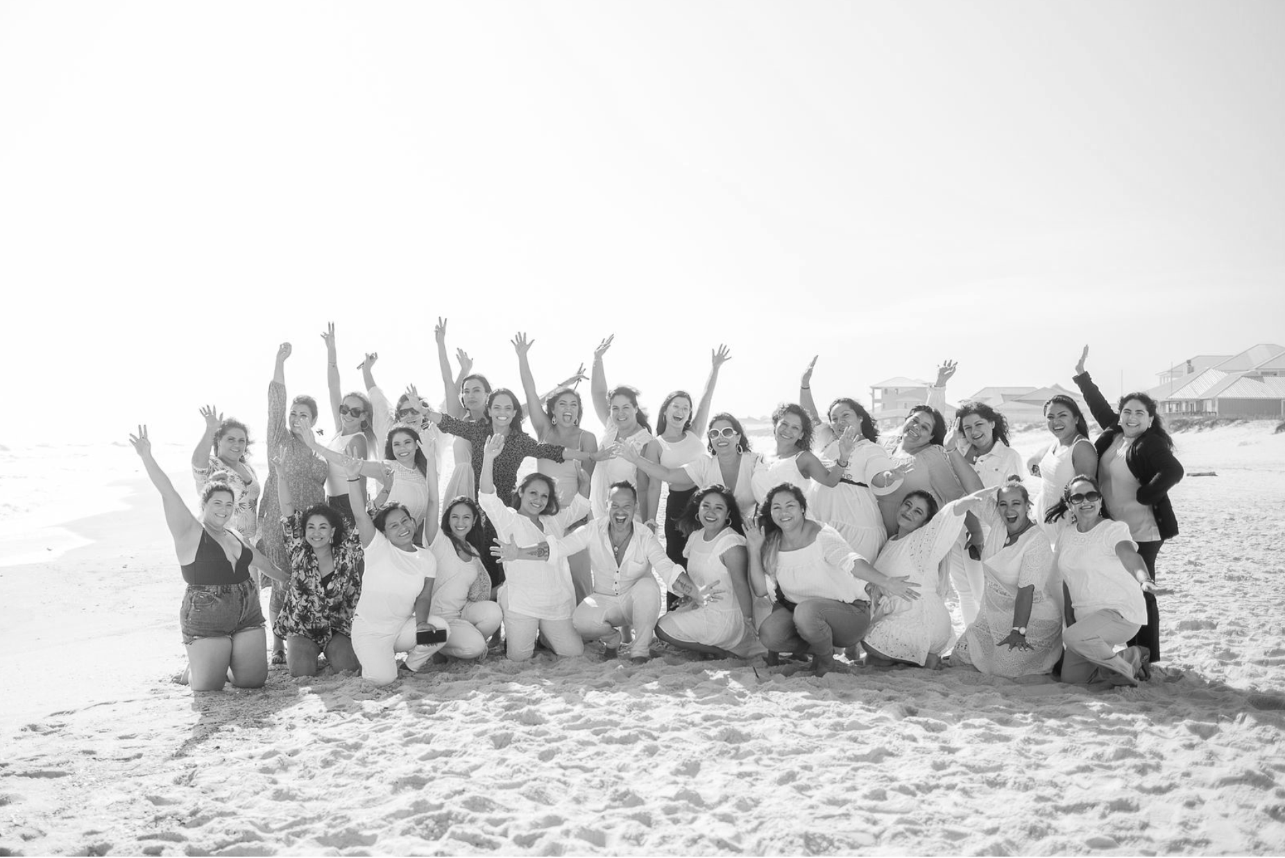 Grupo de mujeres en la playa sonriendo y celebrando, algunas arrodilladas y otras de pie, con fondo de casas y el mar en un día soleado.