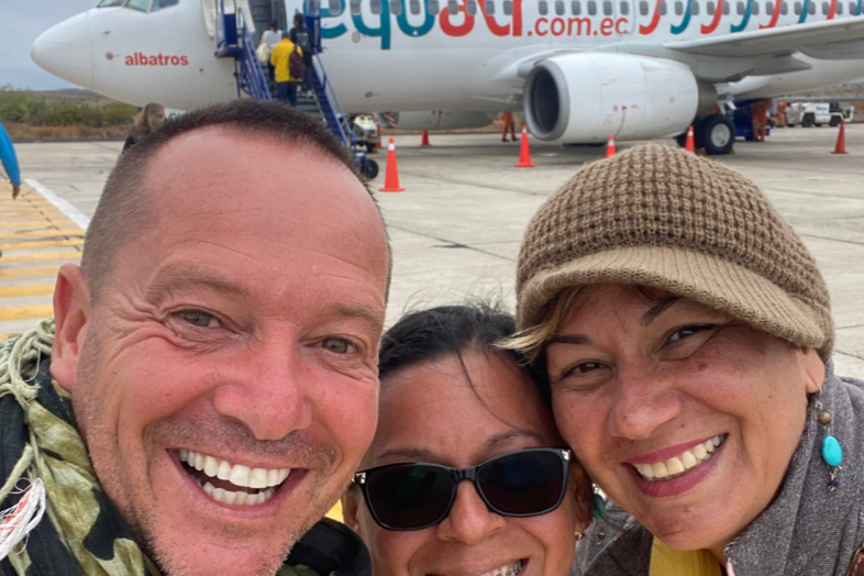 Tres personas sonriendo en un aeropuerto con un avión en el fondo, en la pista de aterrizaje.