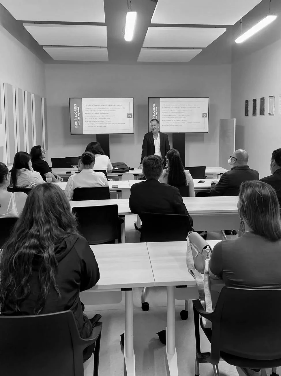 Una sala de conferencias con varias personas sentadas, un presentador de pie frente a dos pantallas con diapositivas, en un entorno profesional.