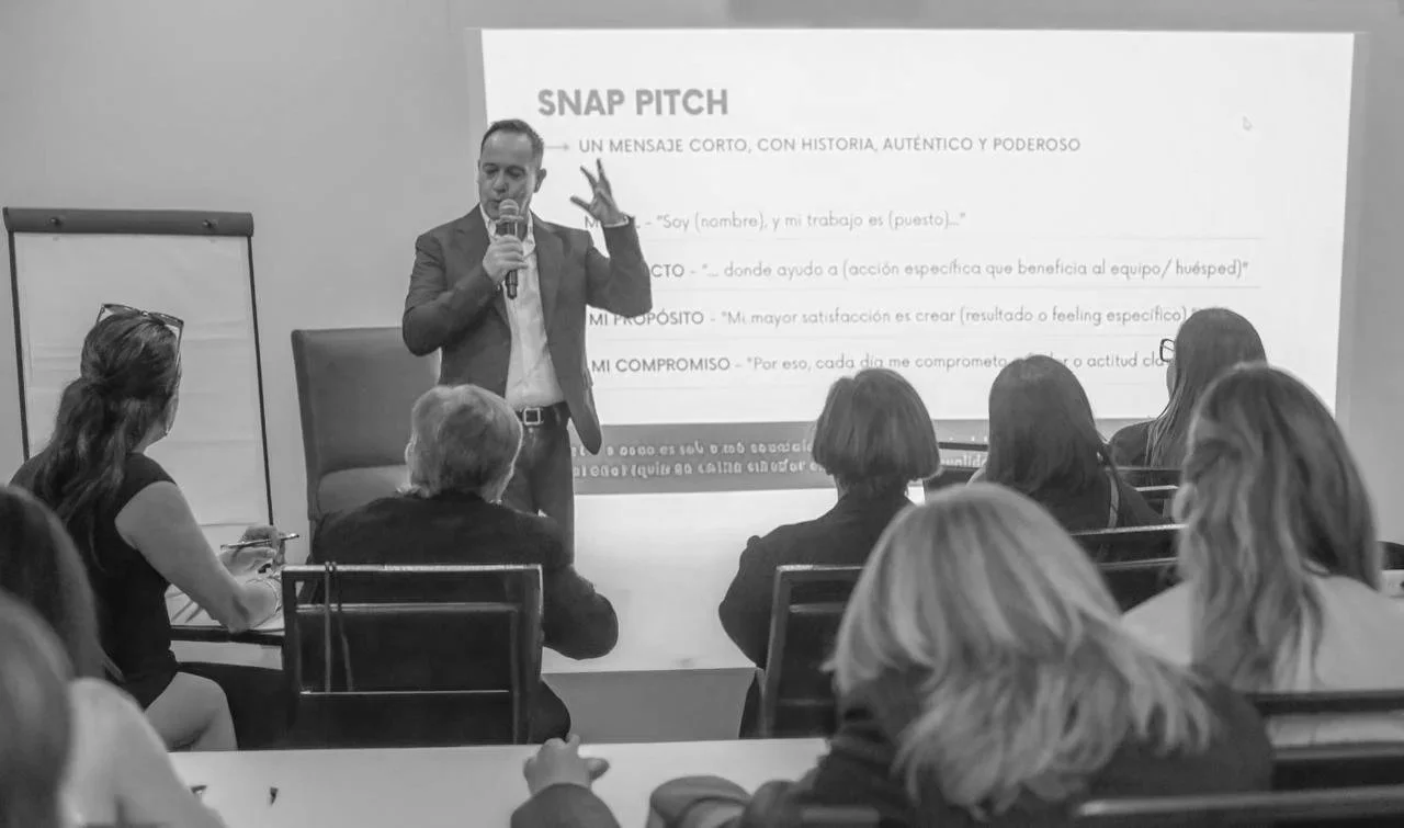 Hombre dando una charla frente a una pantalla que dice 'SNAP PITCH' y varias frases relacionadas con presentación y objetivos, con varias personas sentadas escuchando en una sala de conferencias