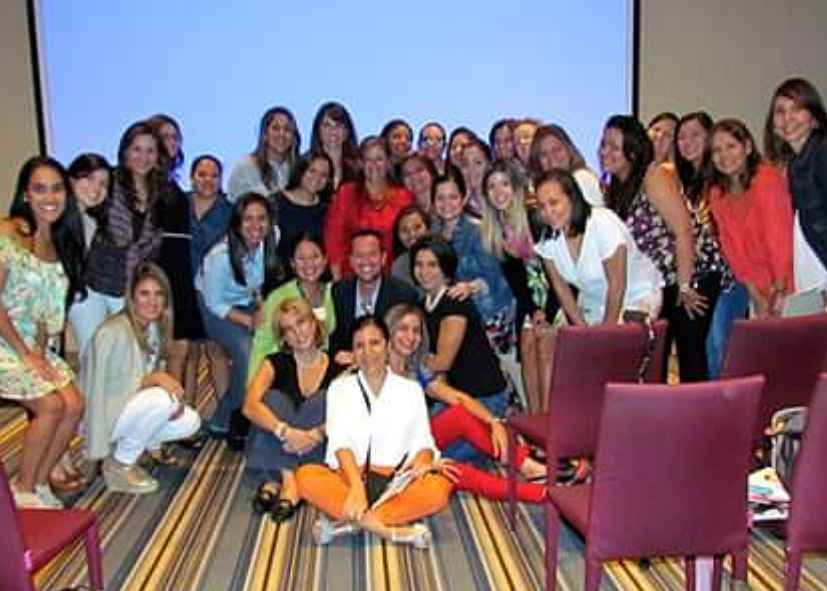 Grupo de mujeres y algunos hombres posando para una foto en una sala de conferencias con sillones rojos y piso con alfombra de rayas.