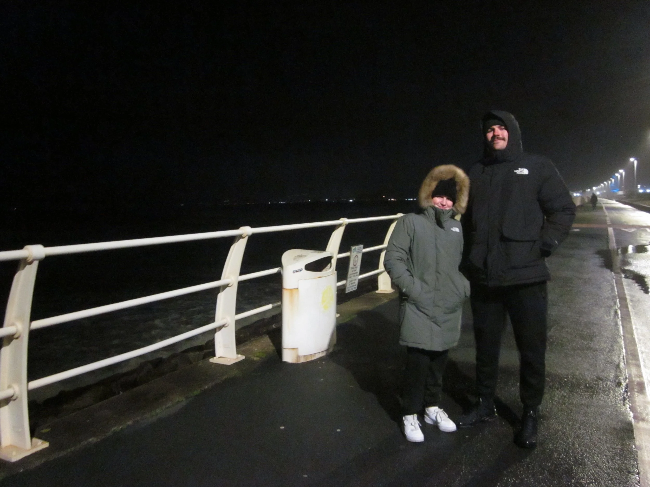 Two people bundled in winter coats standing on a dark pier near the water at night.