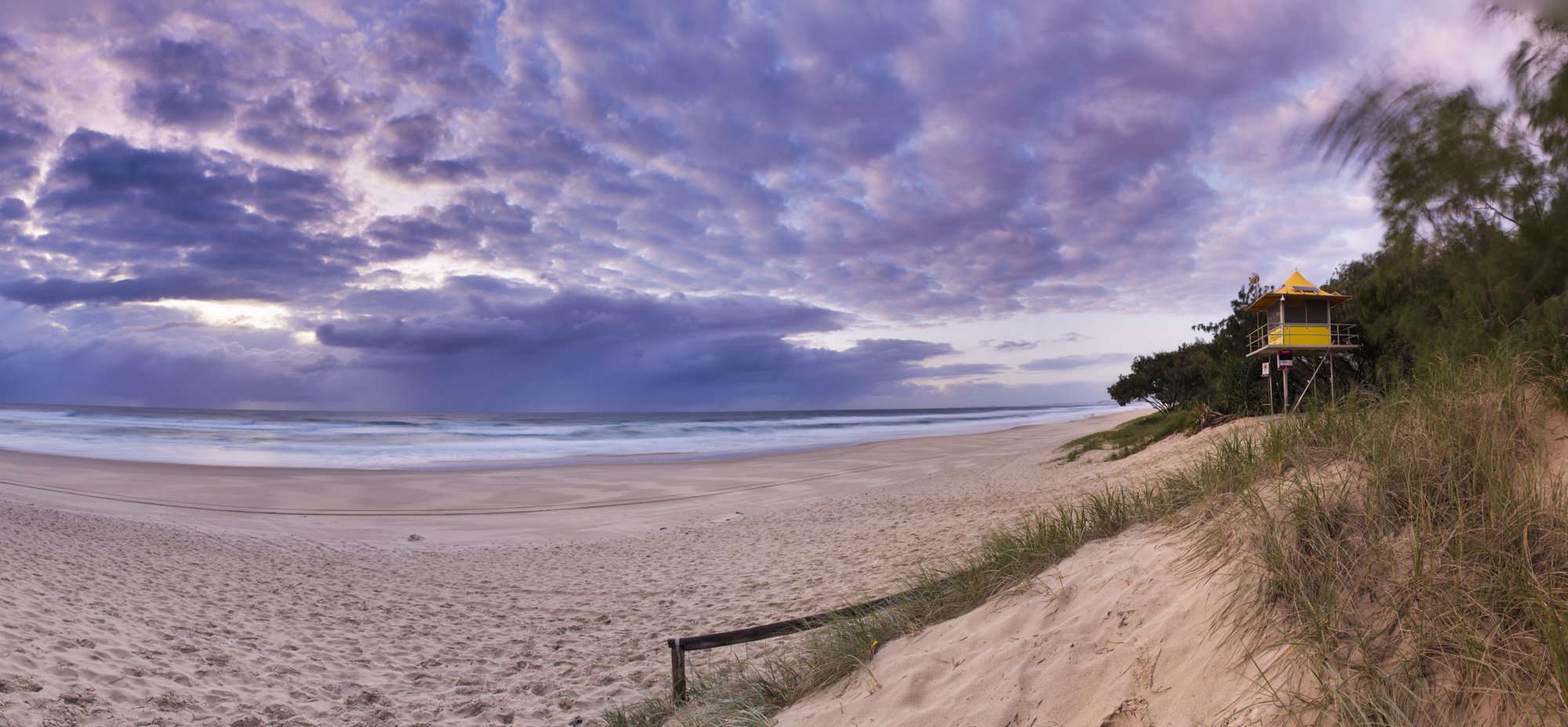 A beach scene at sunset with a sandy shoreline, ocean waves, and a colorful sky with purple and pink clouds. There is a yellow lifeguard tower on the right side near some trees and dune grass.