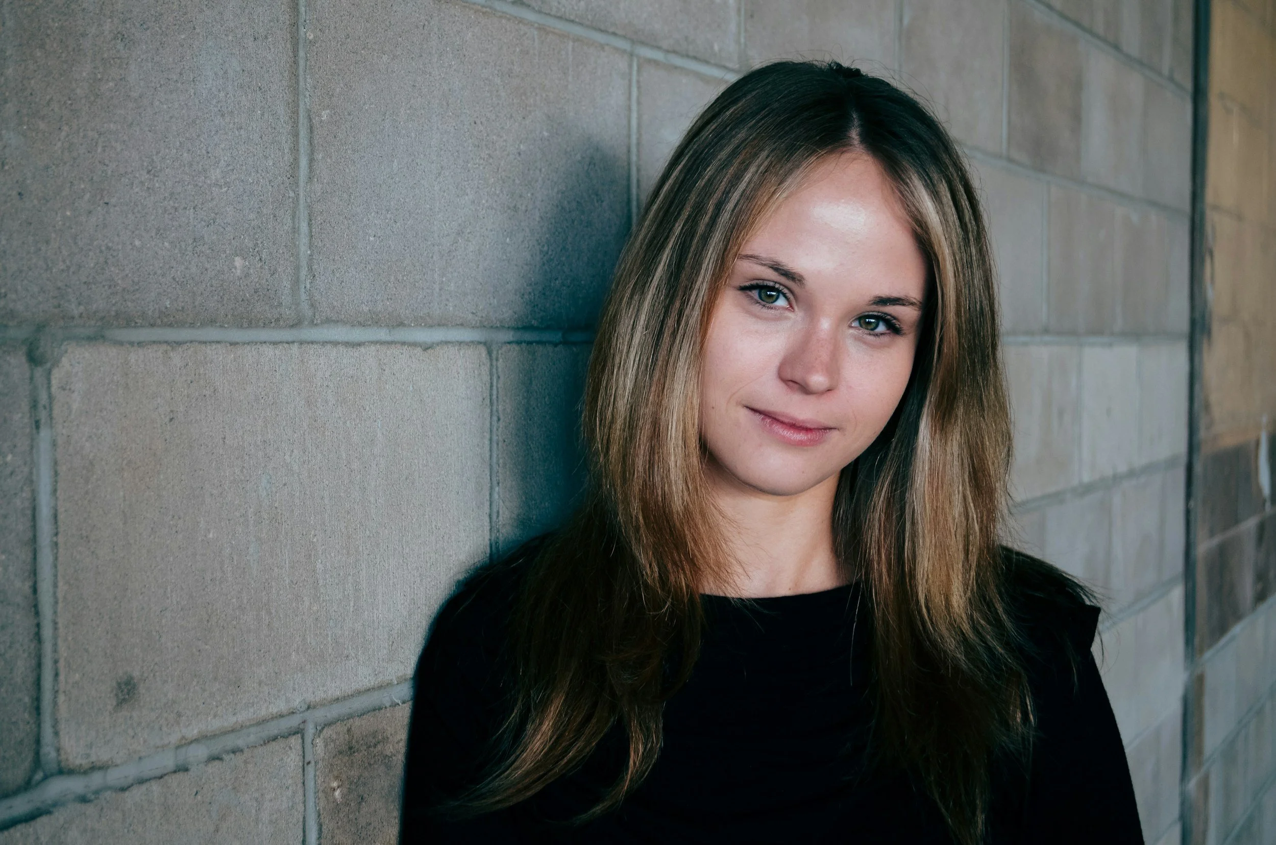 A young HEAL employee woman with long brown hair and blue eyes leaning against a brick wall.
