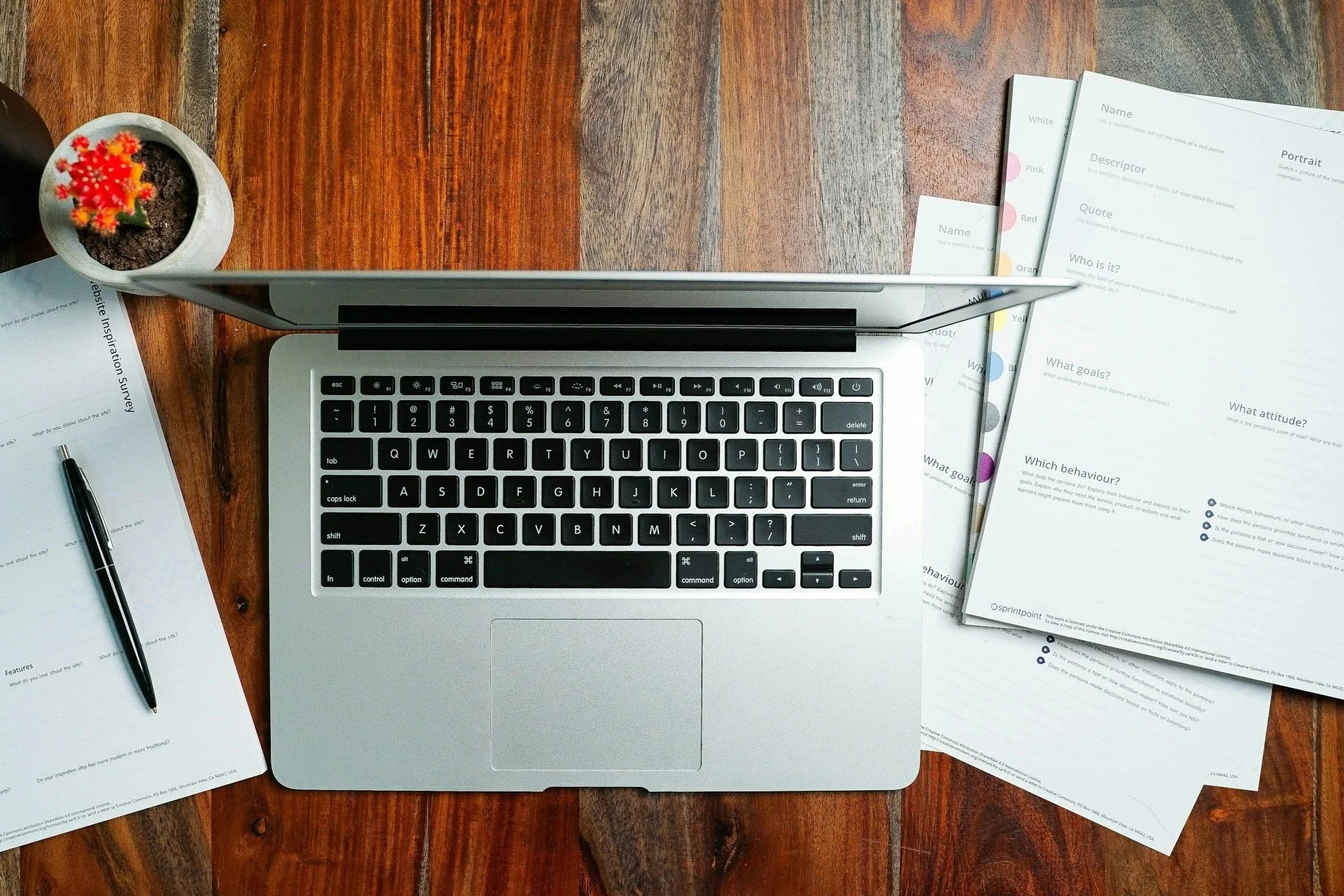 Open laptop on a wooden desk surrounded by papers, a pen, and a small potted cactus.
