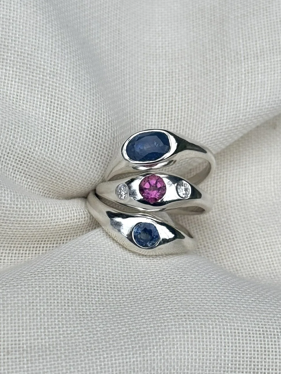 Three bespoke unique silver bombe rings with sapphires, pink tourmalines and diamonds, displayed on white fabric.