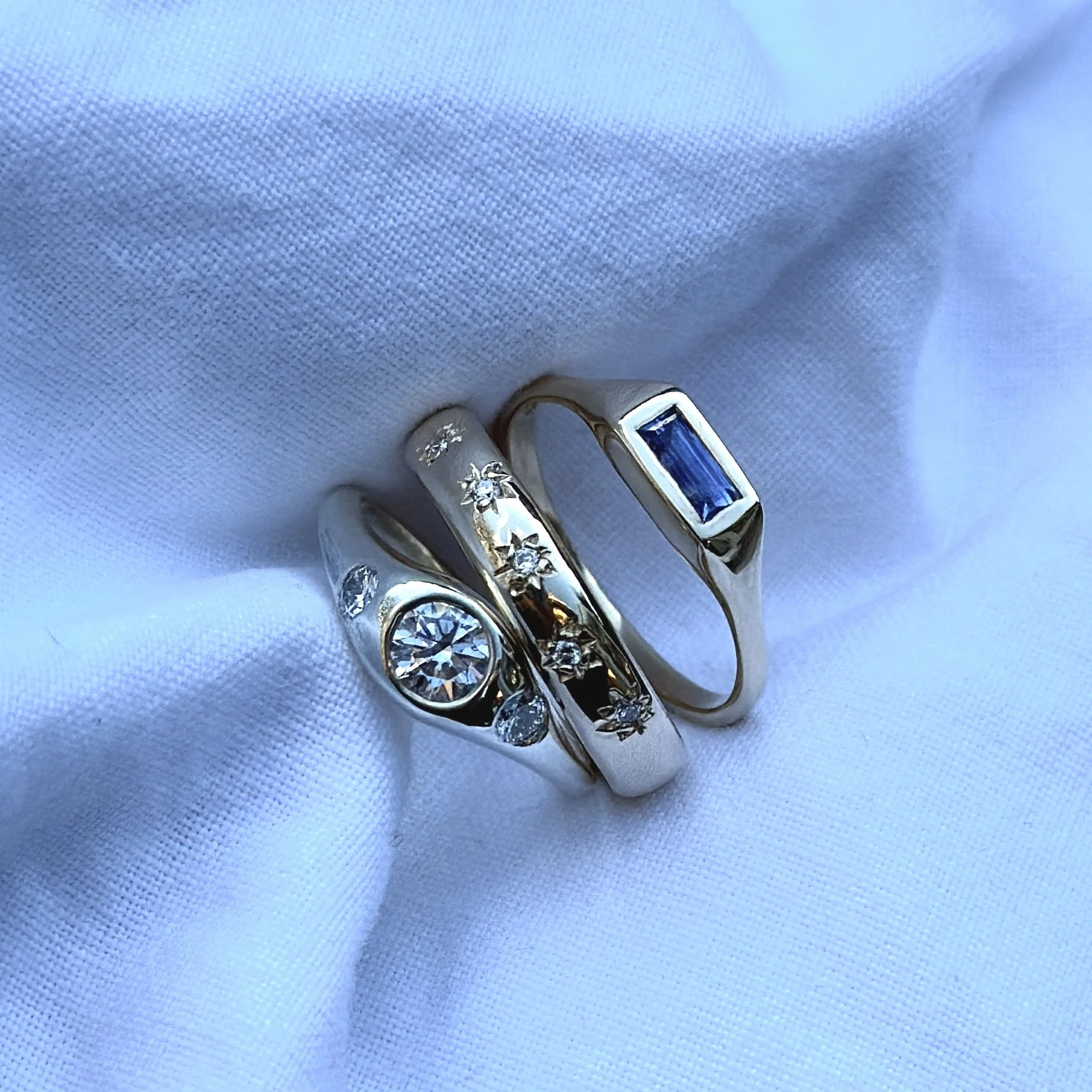 Three 9ct gold rings on white fabric, one with a large round diamond, another with small diamonds and star designs, and the third with a rectangular blue gemstone.