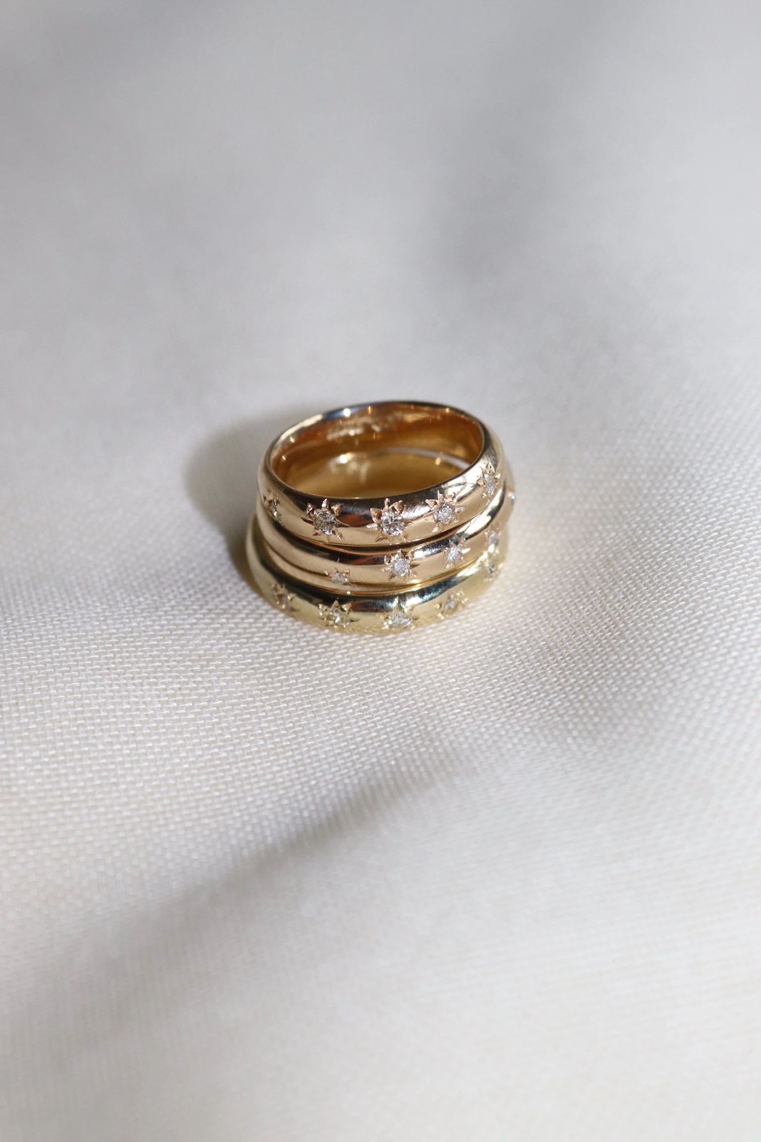 Close-up of three stacked gold rings with embedded small diamonds, placed on a white textured fabric background. Made from heirloom jewellery, melted down and cast into bands. The heirloom stones were then set in to the band.