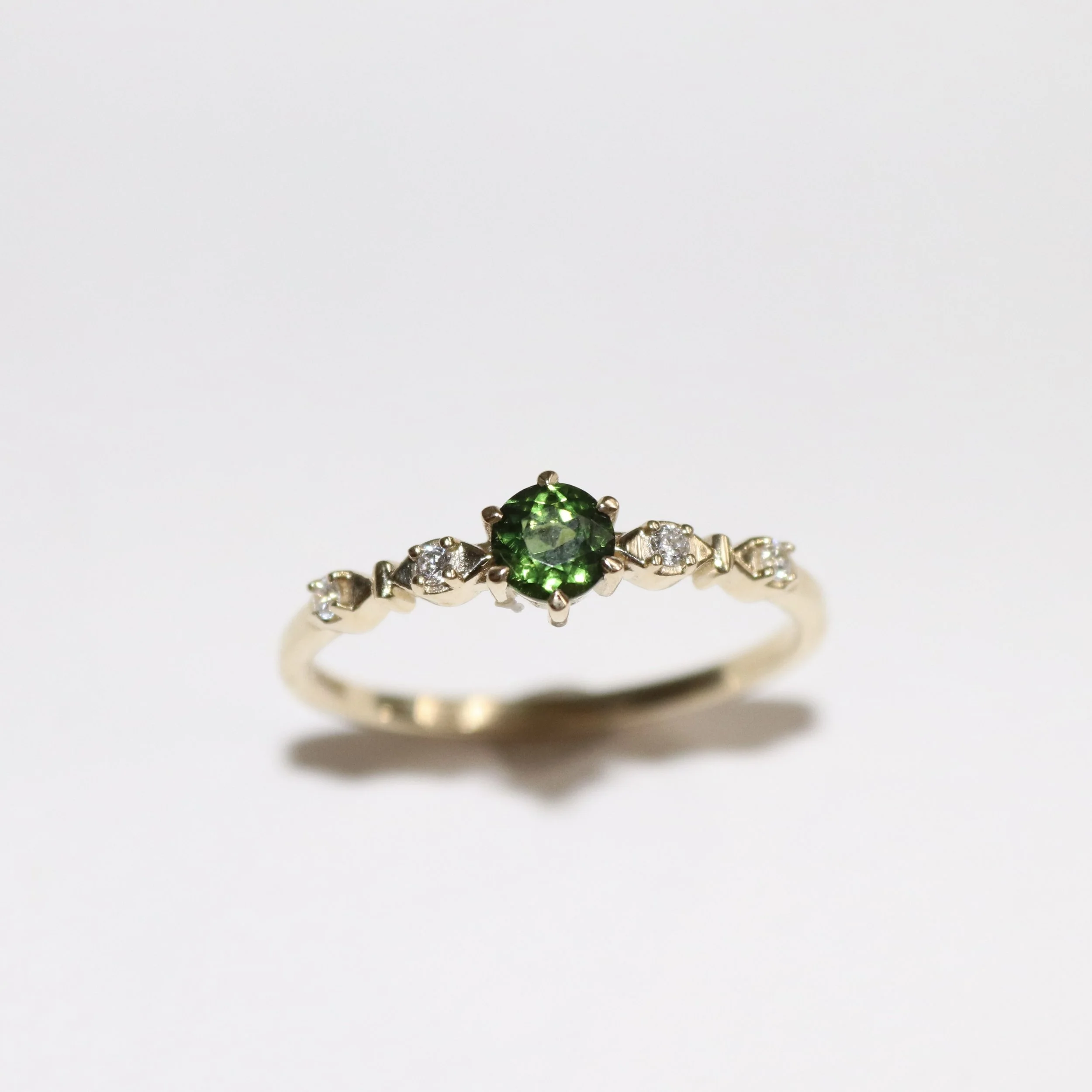 Close-up of a 9ct gold bespoke engagement ring with a large tsavorite gemstone in the cente, surrounded by small clear gemstones, on a white background.