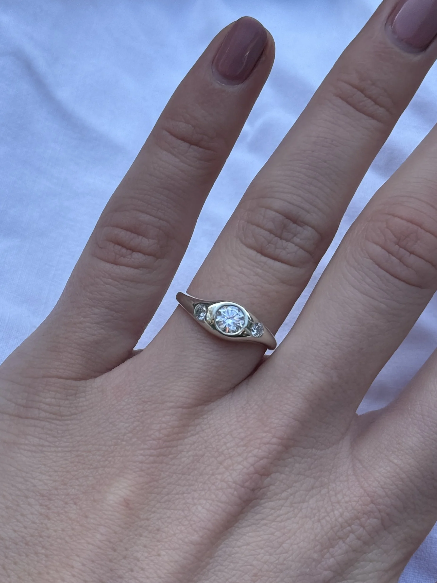 A hand with a silver ring featuring a large central diamond engagement ring and two smaller gemstones on either side, resting on a light-colored fabric background.