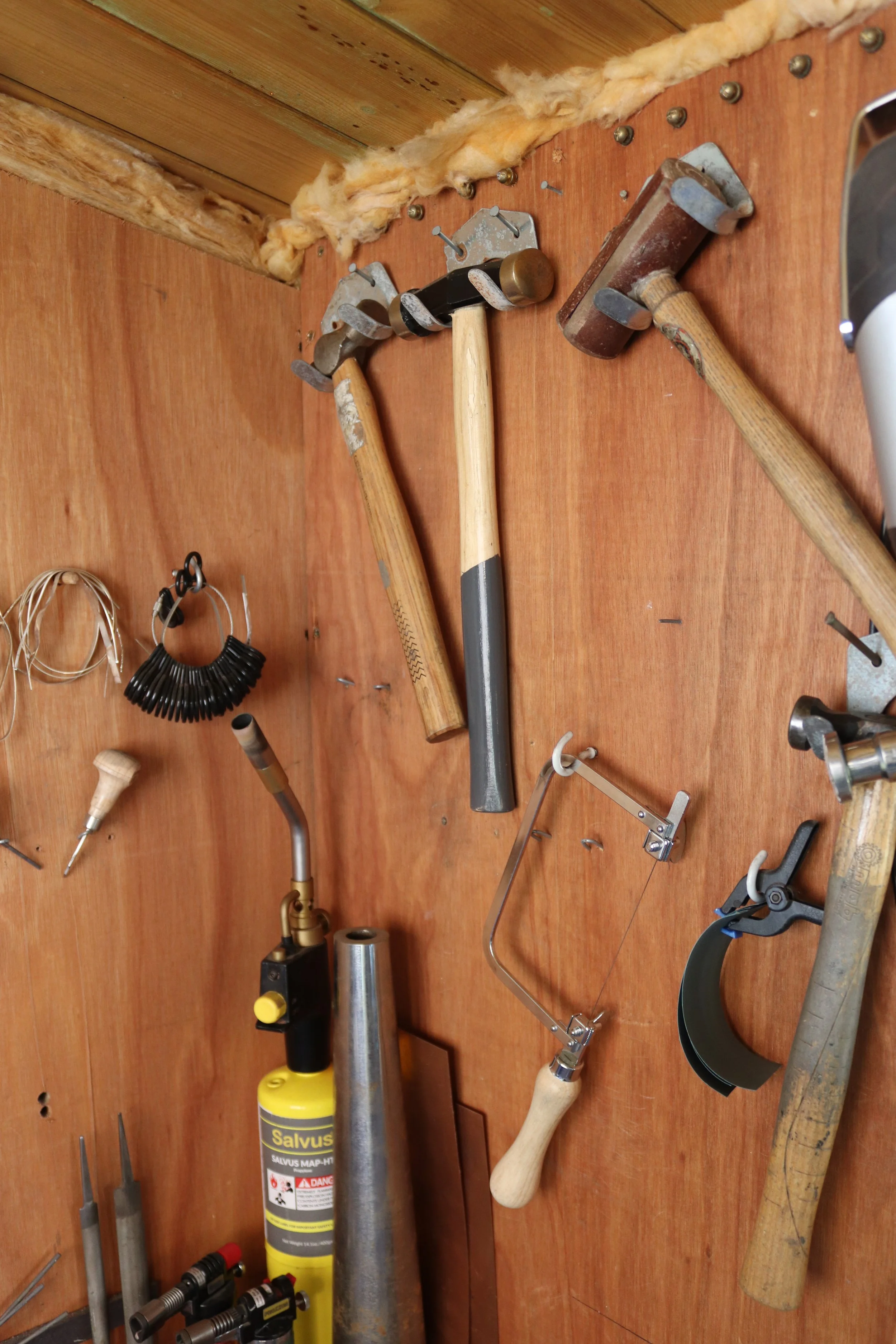 A variety of jewellers tools hanging on a wooden wall, including hammers, soldering tools, blow torches and jewellery making equiptment.