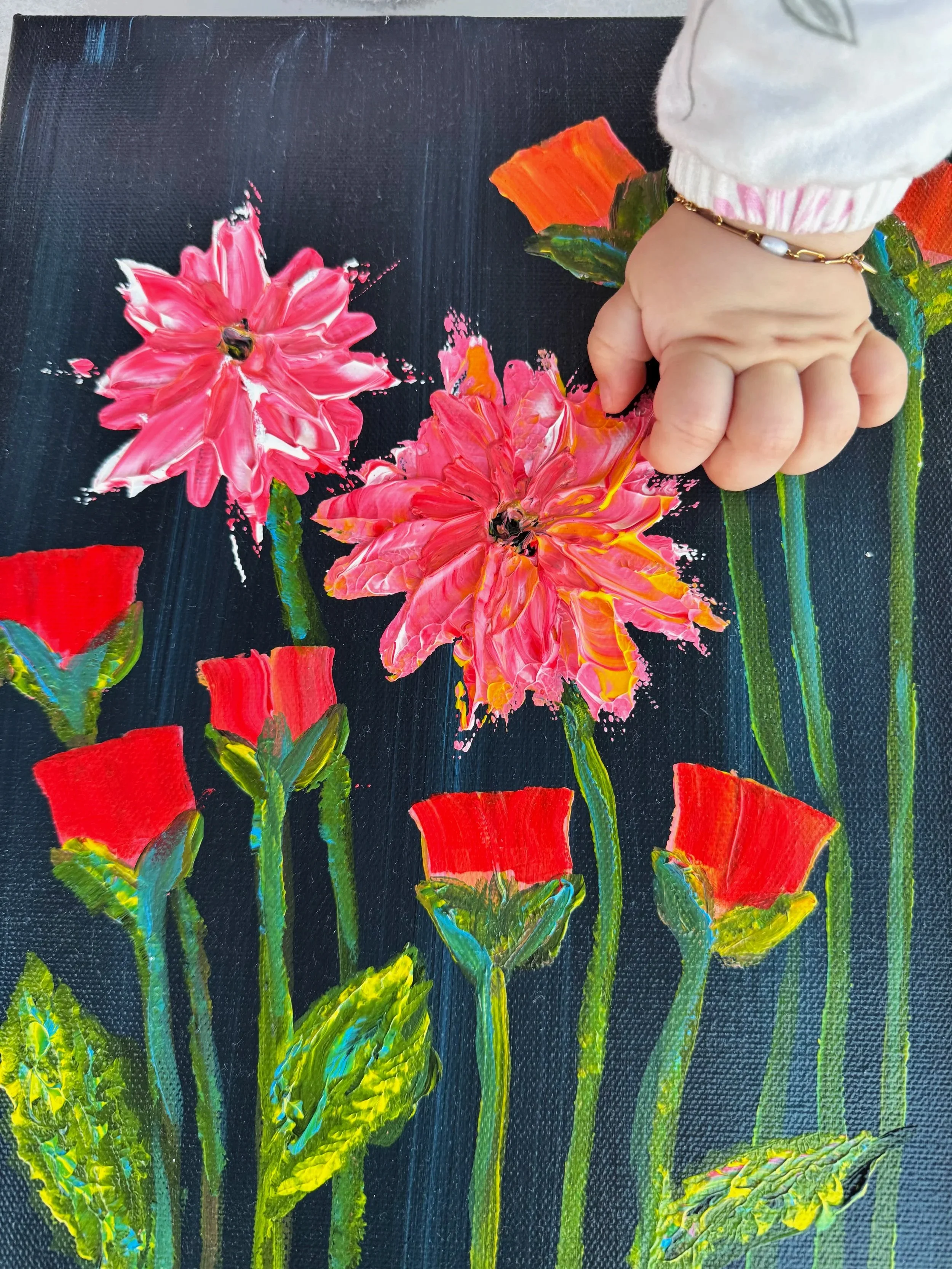 Sophia's hand reaching towards a colorful, textured painting of pink, red, and orange flowers on a dark background painter by her mother the founder of mKunstraum