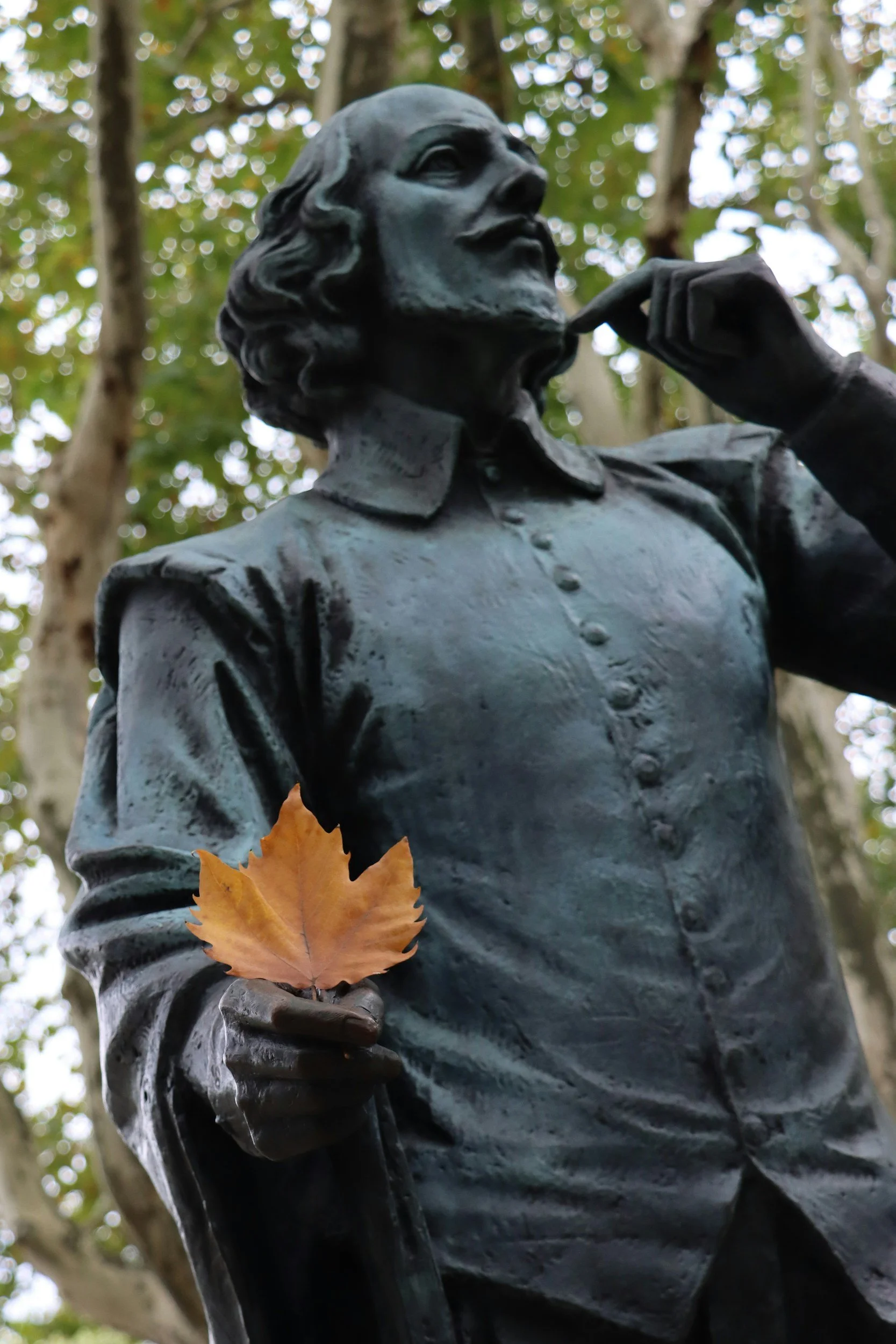 Close-up of a bronze statue of a man with a mustache and long hair, holding an object in one hand and a leaf in the other, against a backdrop of trees with Green leaves and bokeh lighting.