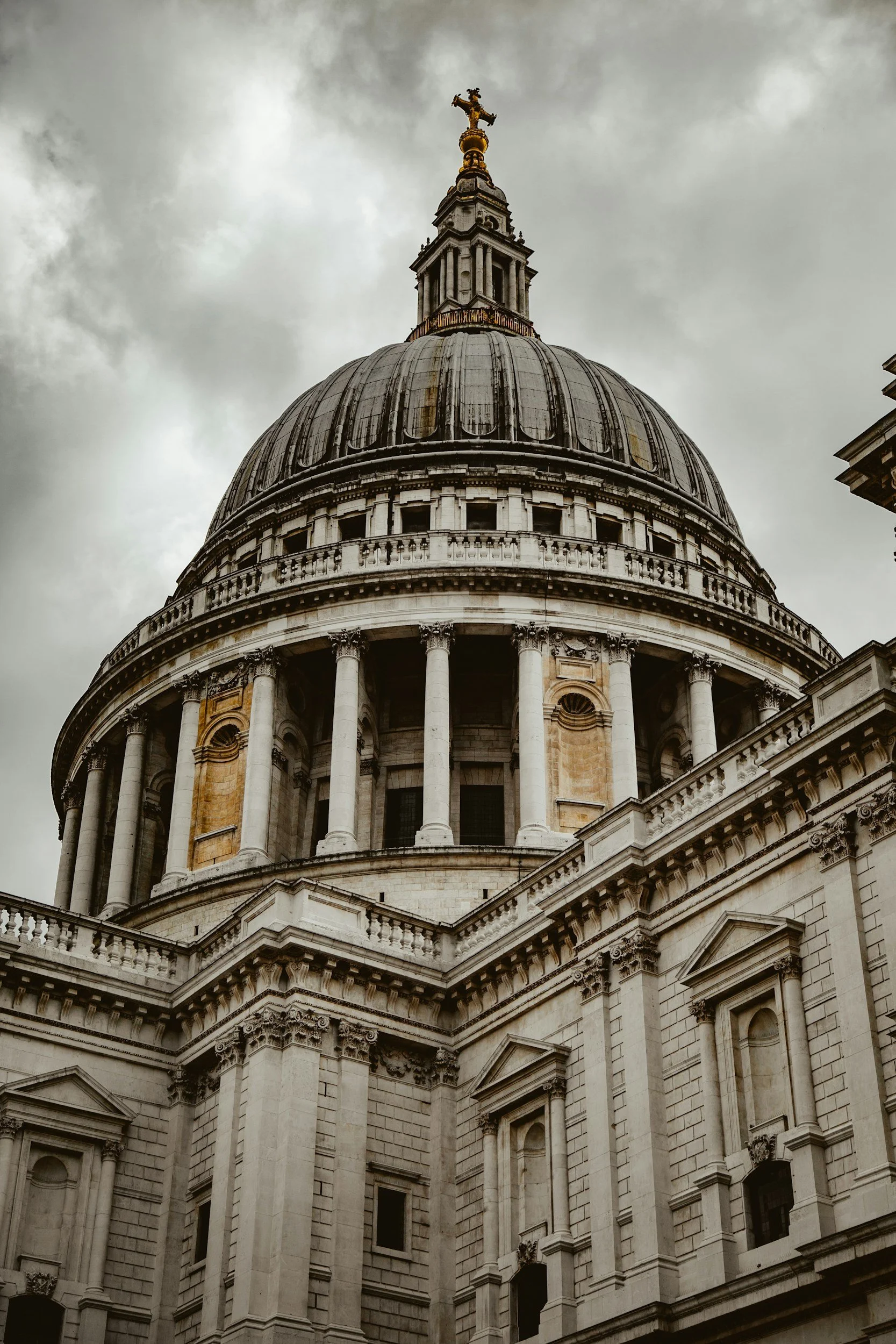The photograph shows the dome and part of the exterior of a historic government building with classical architectural elements, including columns, decorative sculptures, and a statue on top of the dome, against a cloudy sky.