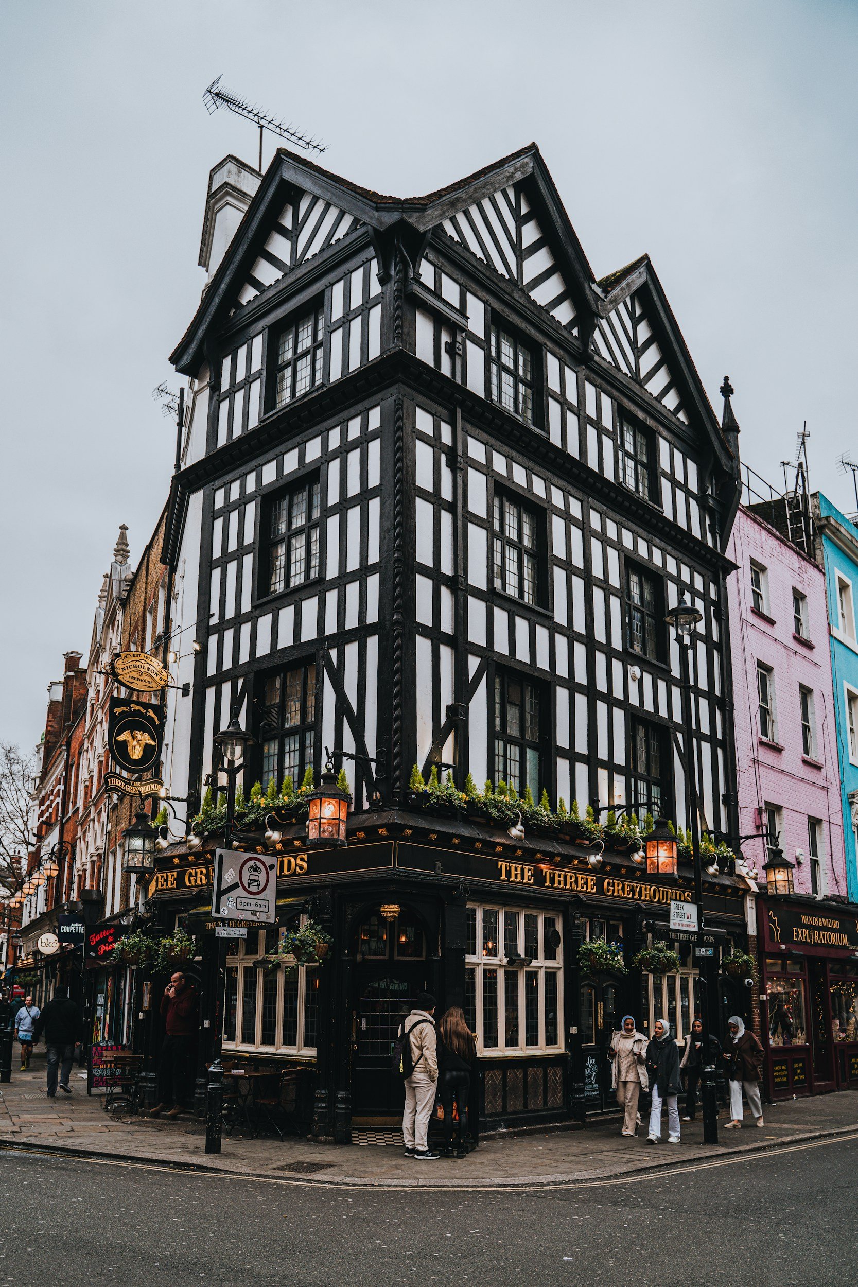 A black and white half-timbered pub on a street corner called 'The Three Greyhounds' with people standing outside and hanging baskets of flowers, in an urban area with other buildings nearby.