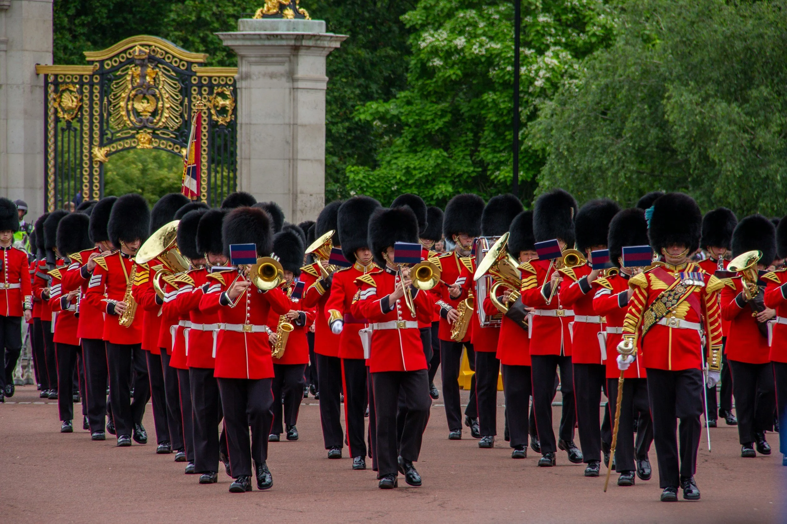 Members of the Queen's Guard in red uniforms and tall bearskin hats marching during a parade at Buckingham Palace with a decorative gate and lush green trees in the background.