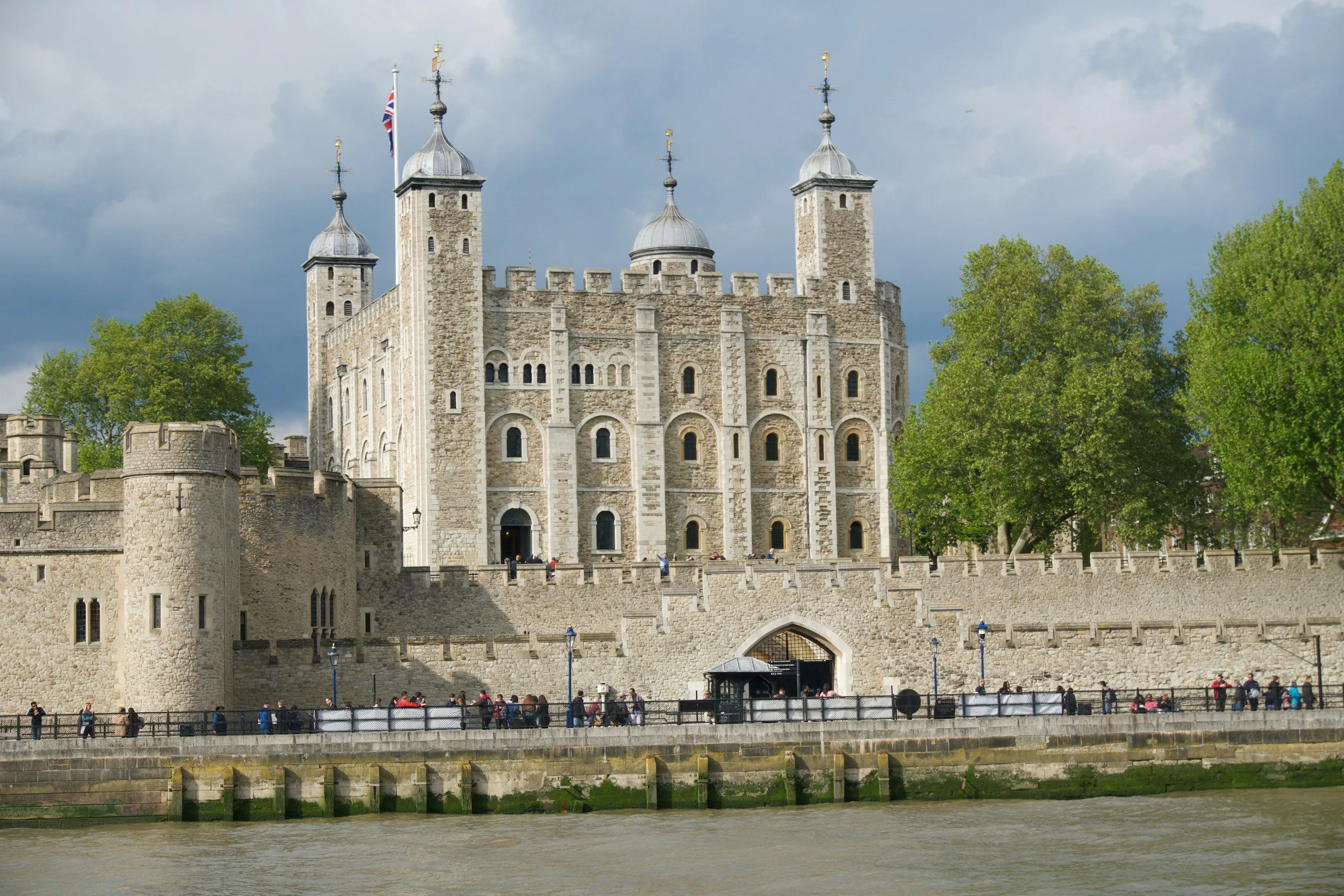 The image depicts the historic Tower of London, a stone castle with four towers, surrounded by a high stone wall, and located next to the River Thames in London, England. The sky is partly cloudy, and there is greenery on the right side of the image. Visitors are seen walking along the pathway in front of the castle.