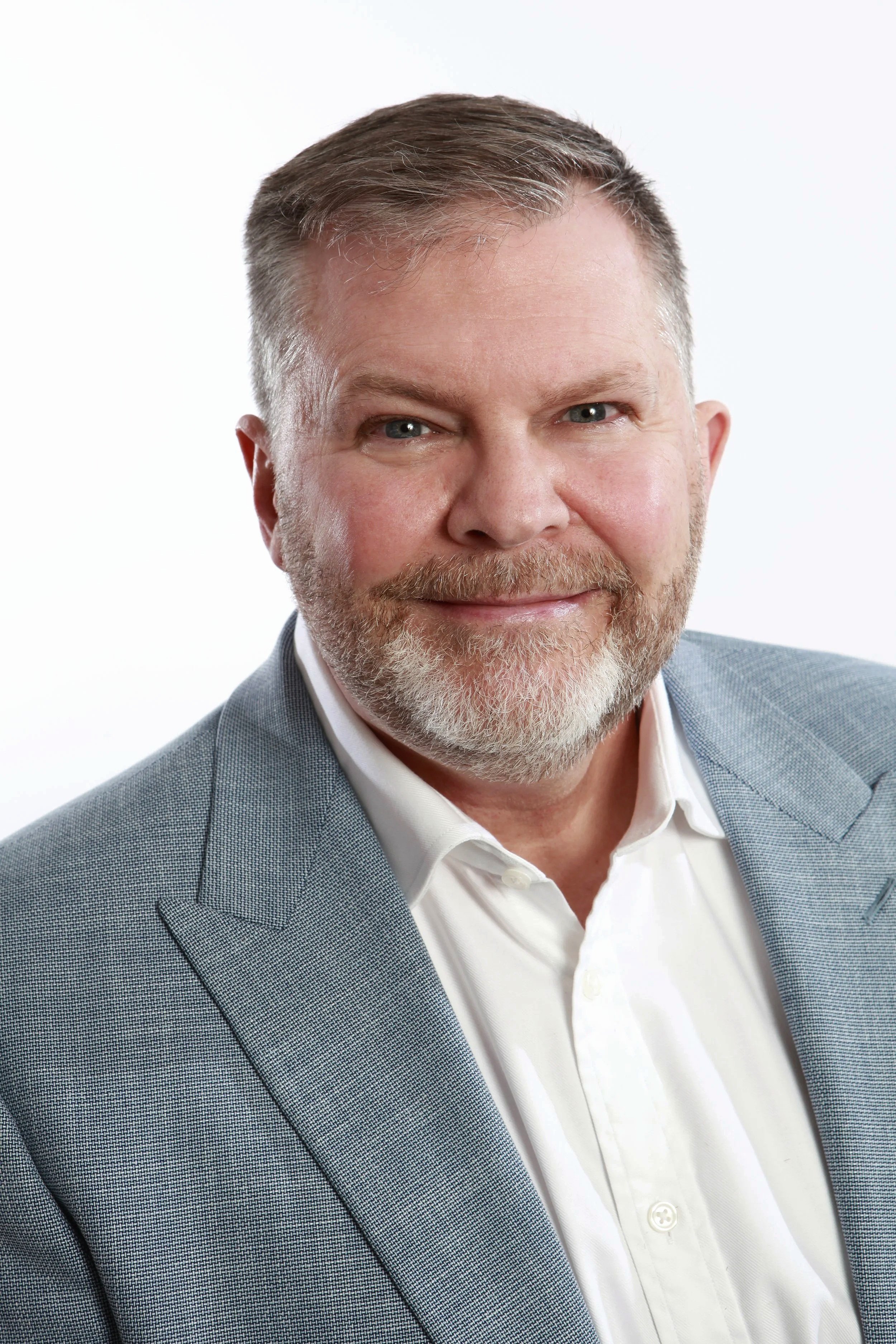 Headshot of a handsome mature man with shiny brown hair, a fashionable stubble beard, and piercing blue eyes, wearing a light gray suit jacket and a white shirt, smiling against a white background.