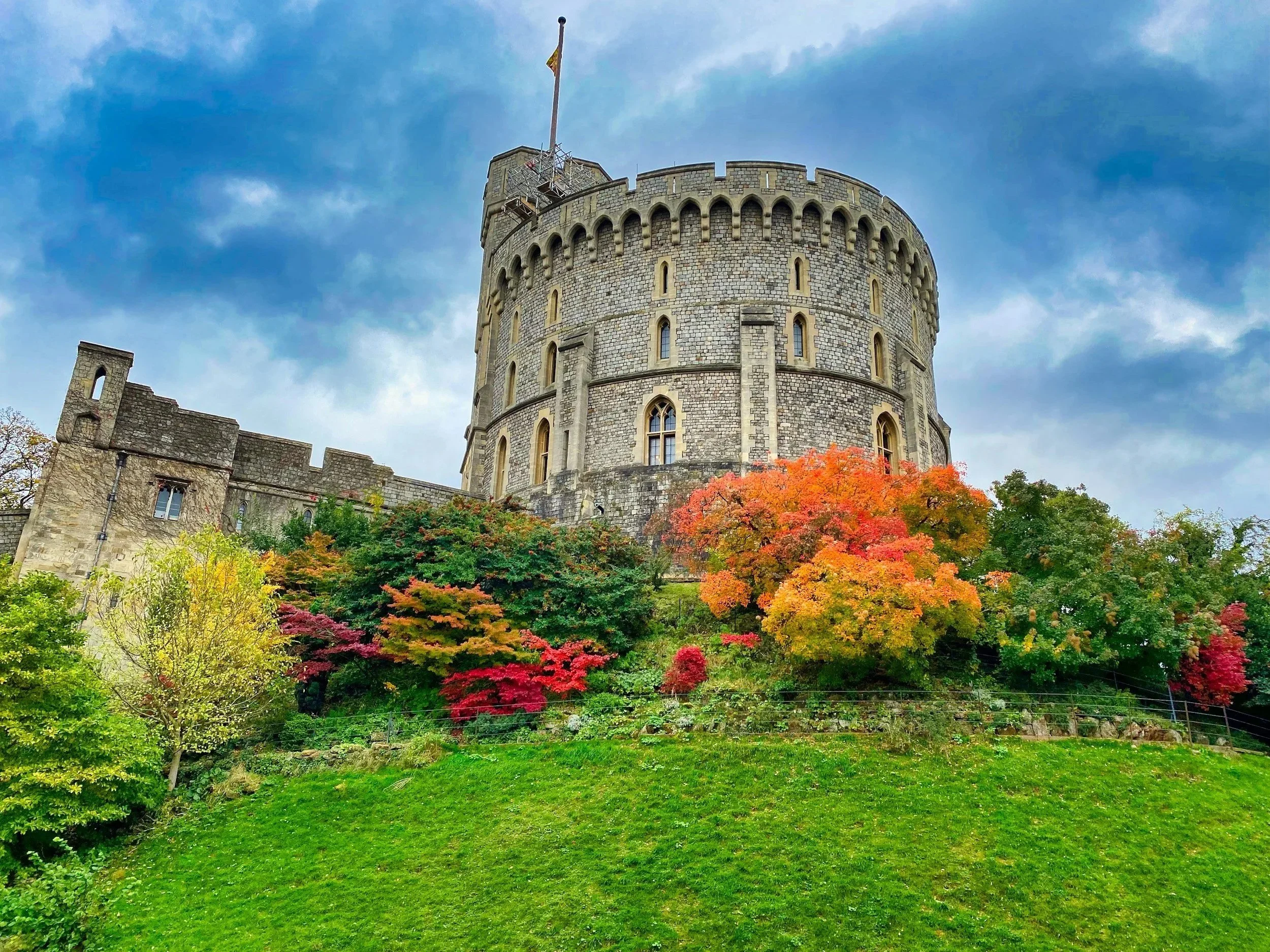 A historic stone castle with a rounded tower featuring narrow windows, surrounded by vibrant autumn foliage with red, orange, and green leaves, under a cloudy blue sky.