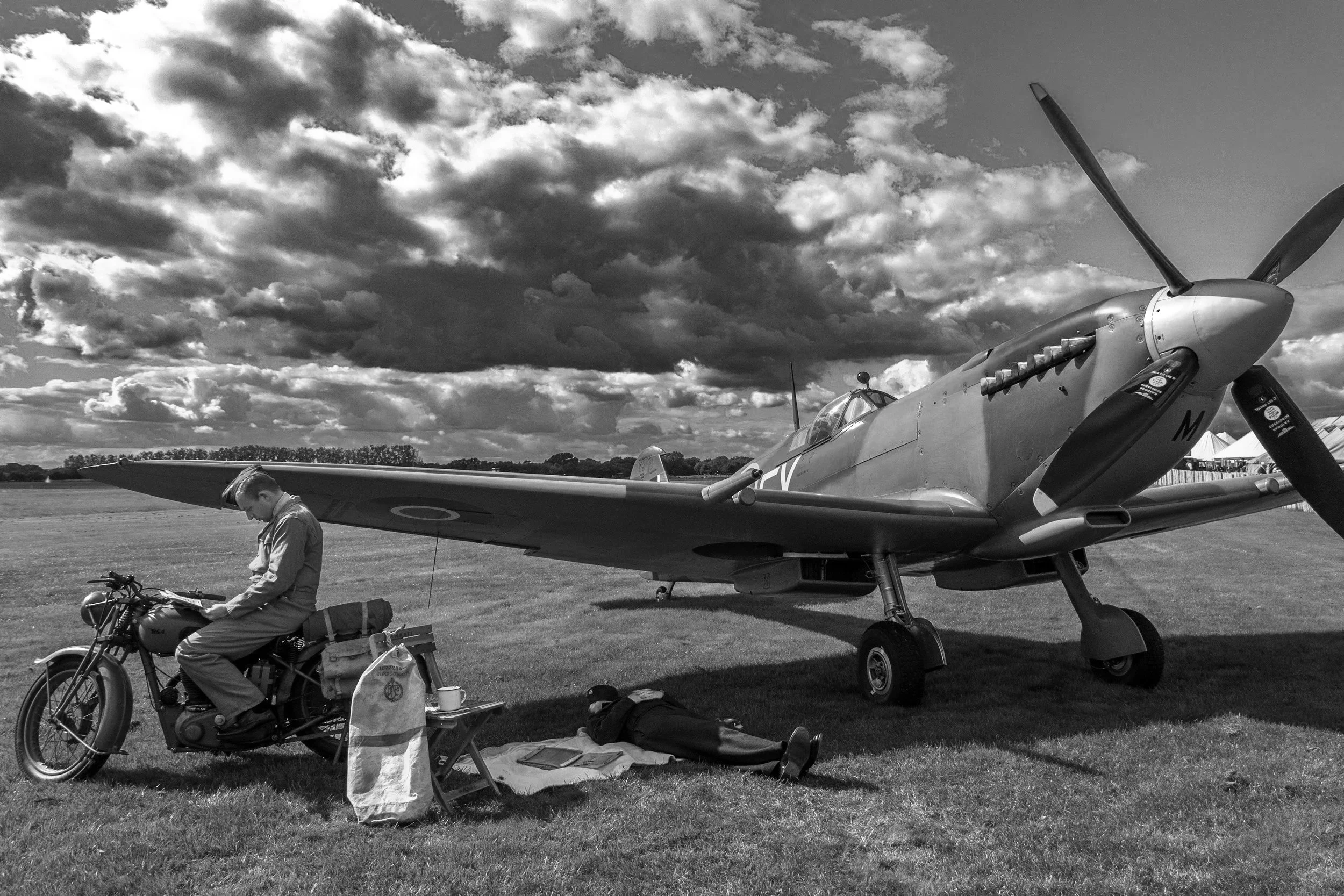 A vintage military aircraft parked on a grassy field under a cloudy sky. A person in military attire is sitting on a motorcycle nearby, with various bags and gear placed on the ground around them.