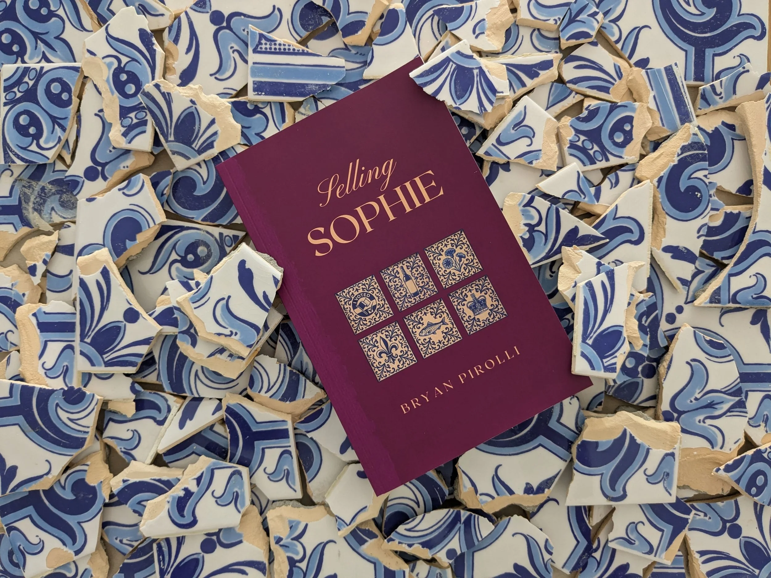 Broken pieces of decorative blue and white ceramic tiles scattered on a surface, with a maroon card titled 'Selling Sophie' placed among them.