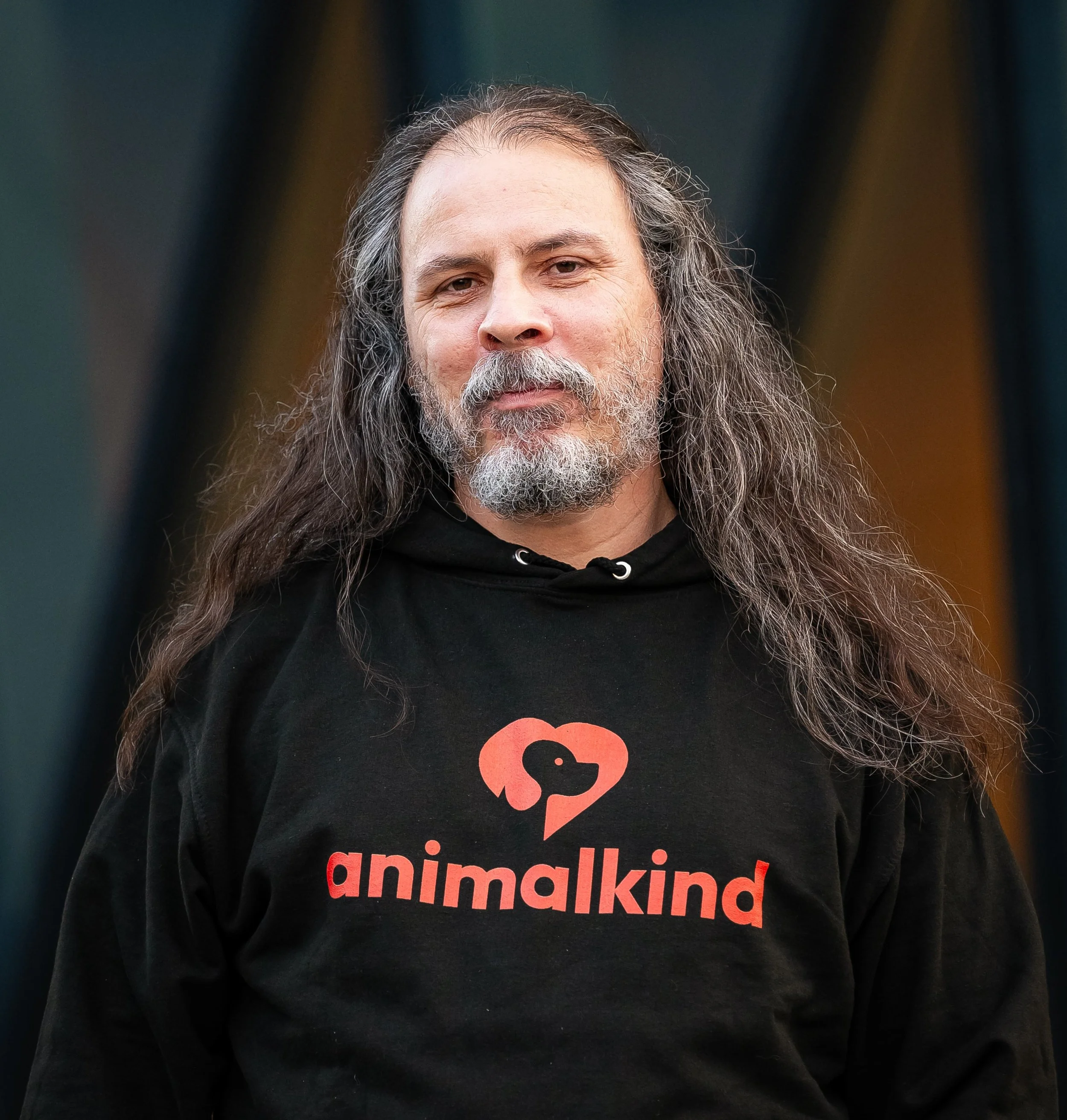 En man med långt hår och skägg bär en svart hoodie och en svart t-shirt med en röd och svart logo av en hunds huvud inuti ett hjärta, samt ordet 'animalkind'.