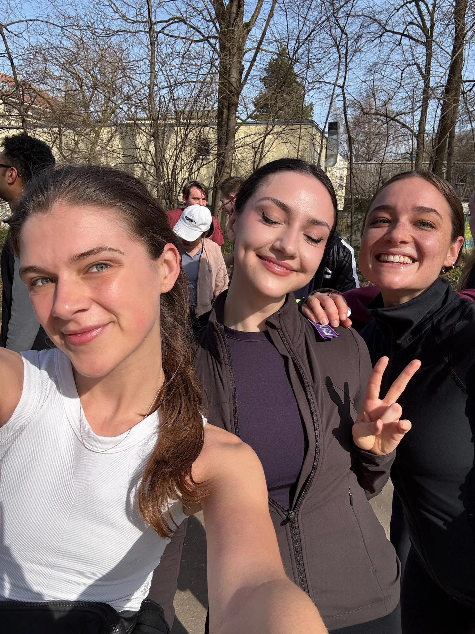 Three young women taking a selfie together outdoors in a park, smiling and making peace signs, with a group of people and trees in the background on a sunny day.