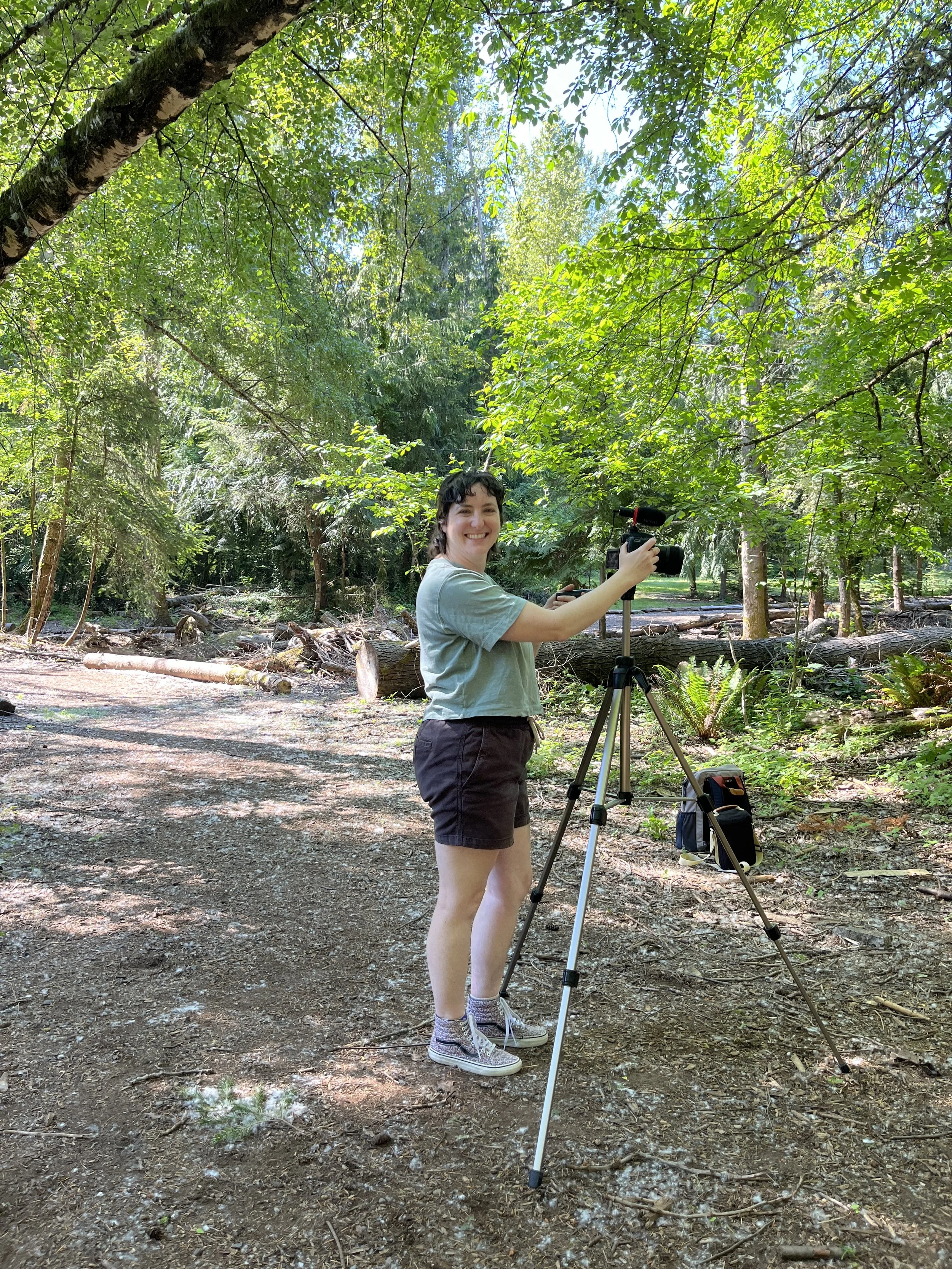 A young woman standing on a dirt trail in a lush green forest, smiling at the camera. She is holding a camera mounted on a tripod and wearing casual clothes including a light-colored t-shirt, black shorts, and sneakers. There are trees and fallen logs in the background.