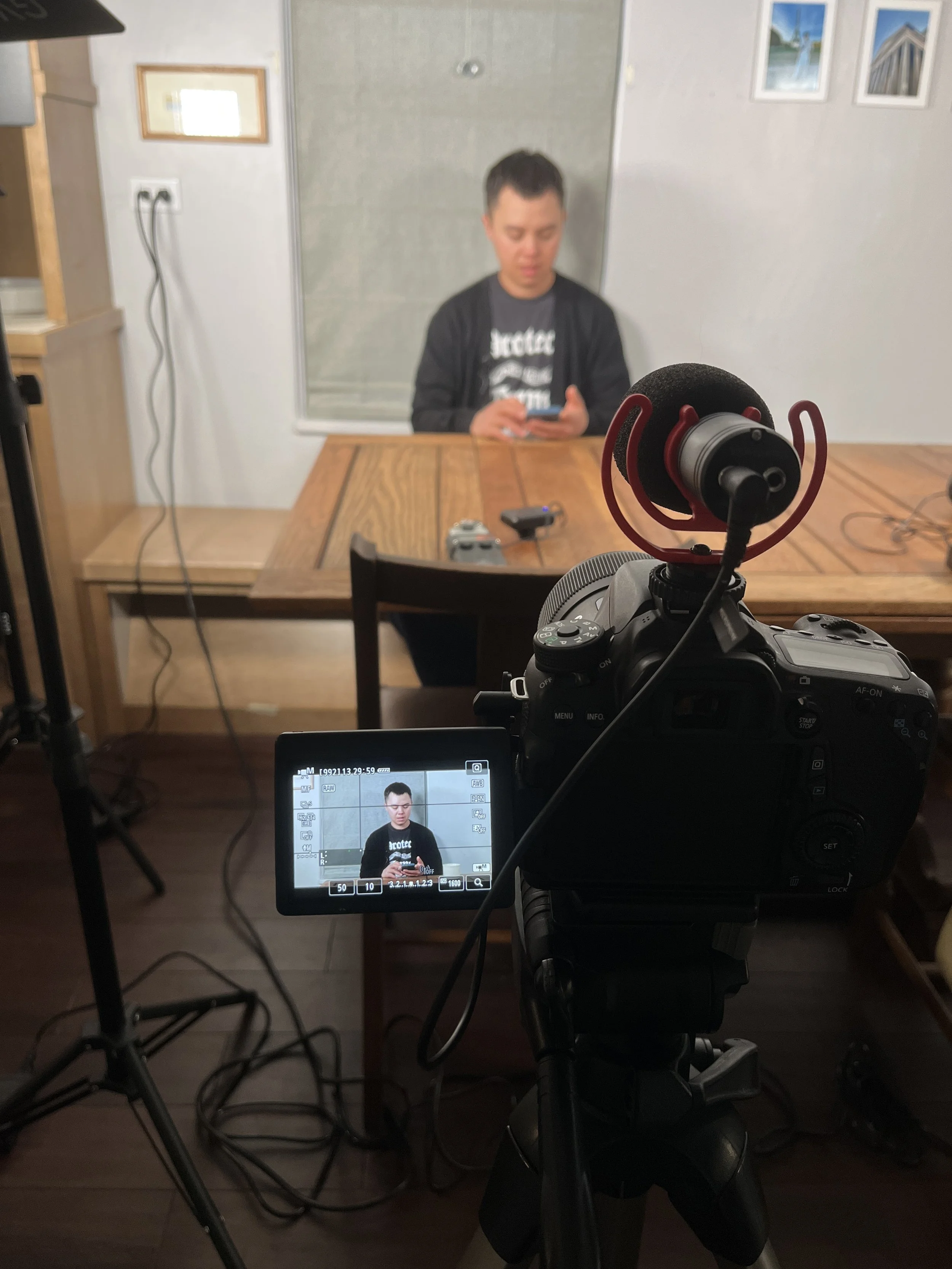 A person sitting at a wooden table on-set with a camera setup recording them. He is looking down at a smartphone. The scene is captured through the view of a professional camera with a microphone mounted on top.