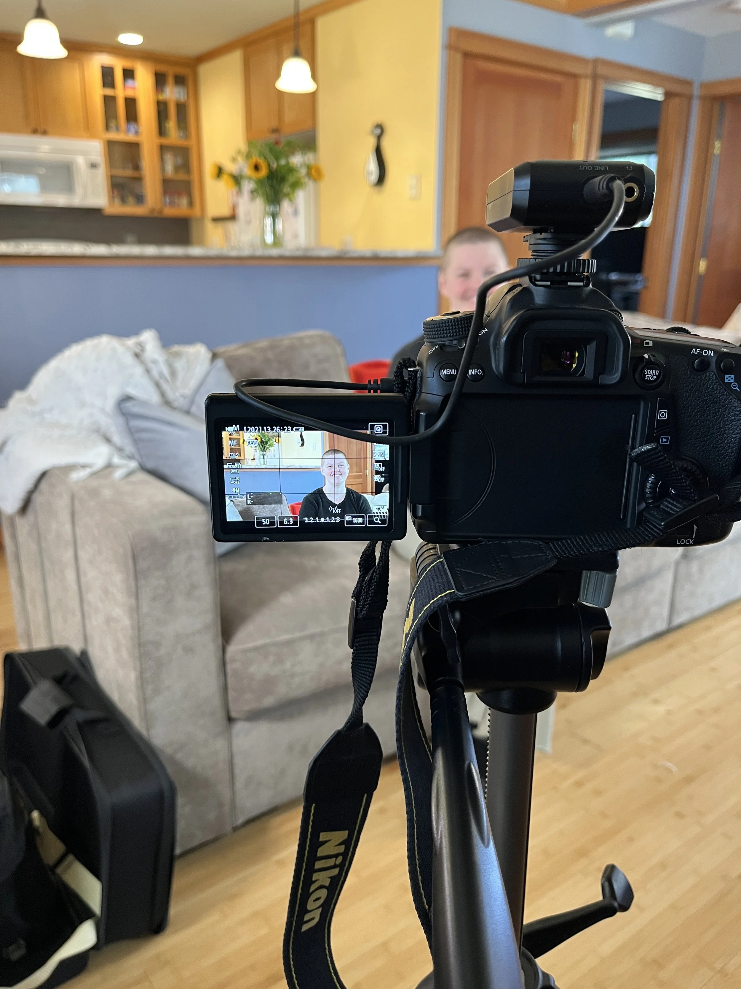 Camera on tripod recording a person on-set sitting on a sofa in a cozy kitchen.
