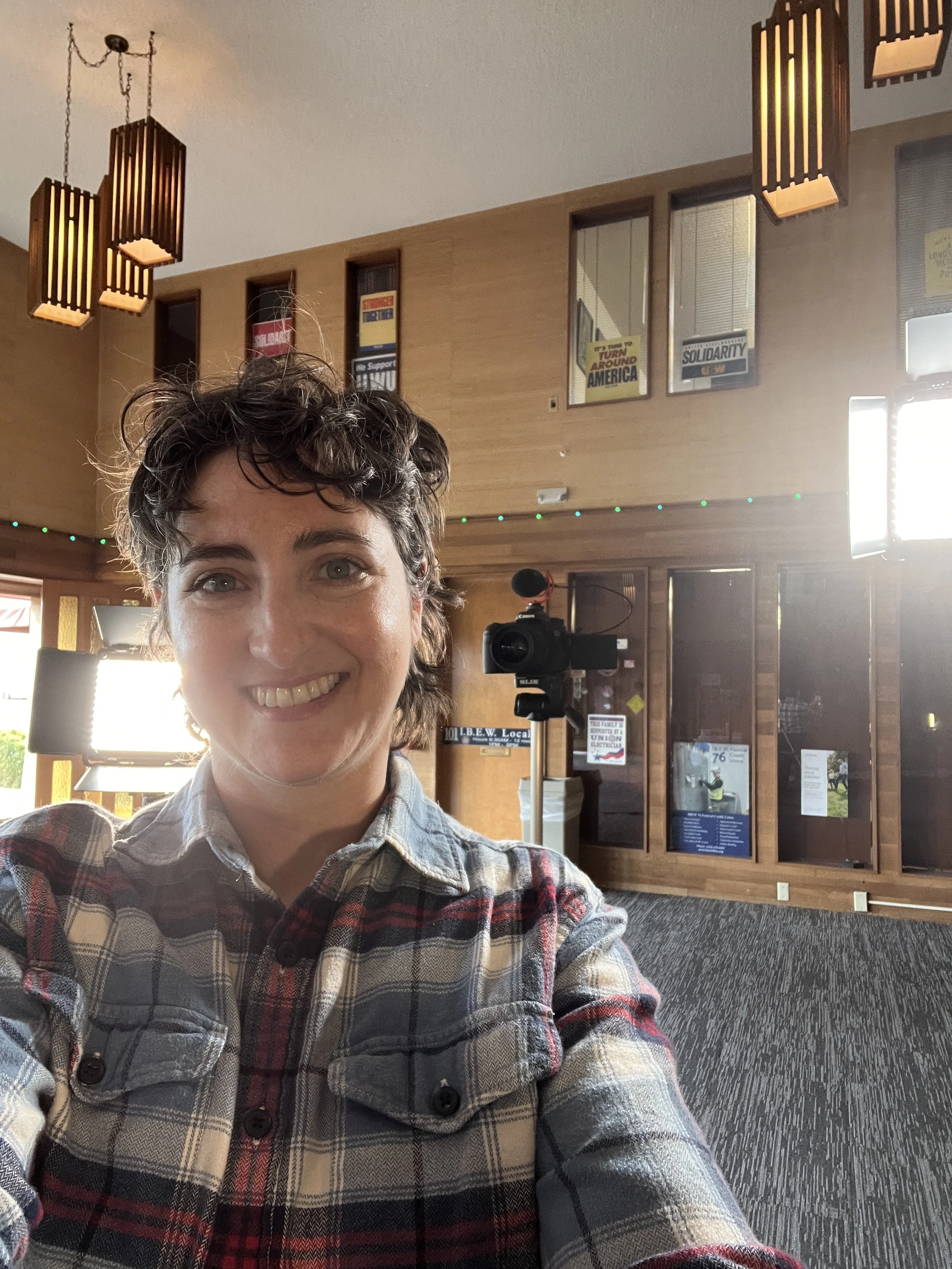 Jen taking a selfie on-set with a smile, wearing a plaid shirt, with a background featuring wooden wall panels, hanging lights, a camera on a tripod, and signs on the wall.
