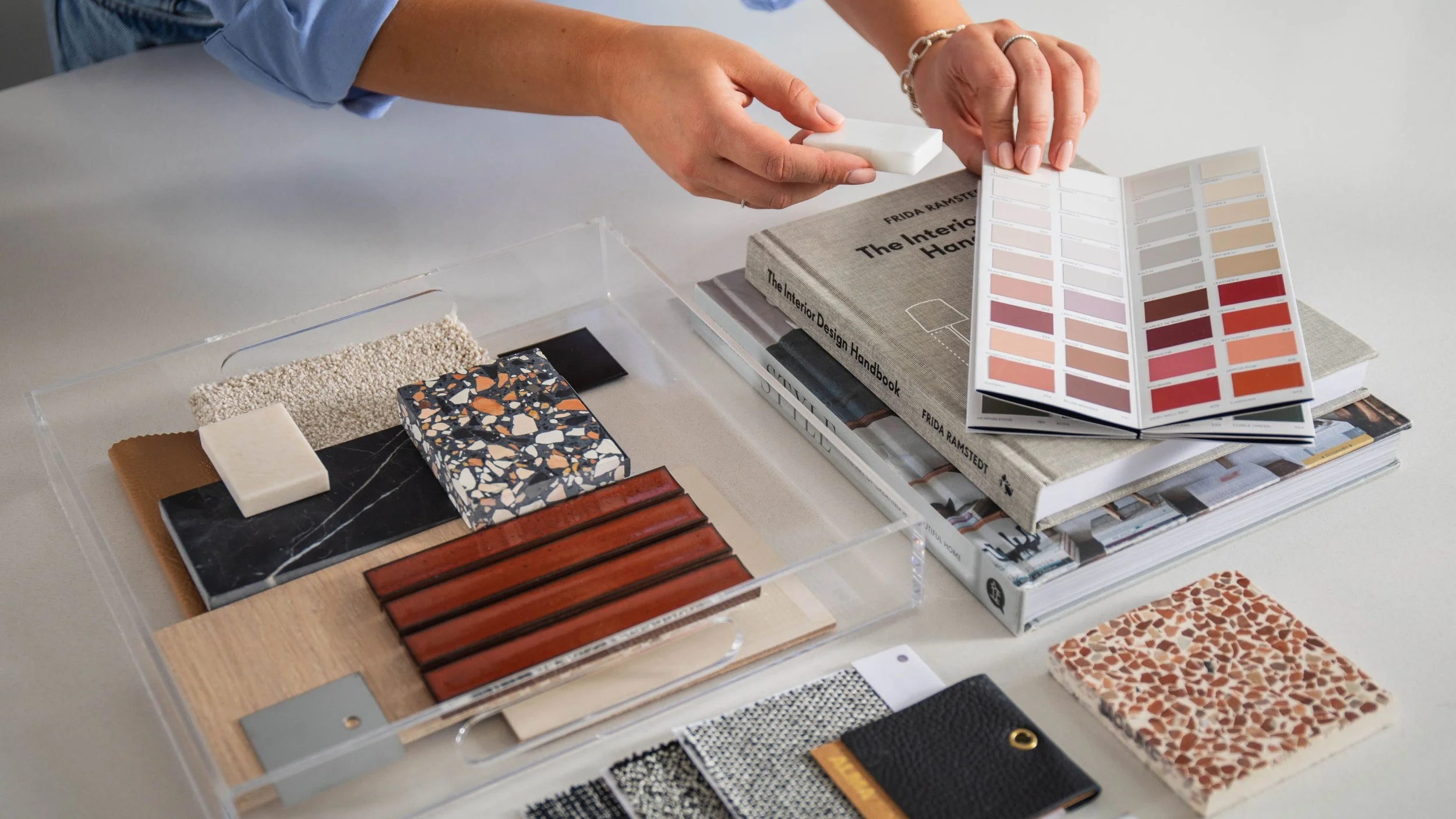 Affordable Interior Designer in London and Surrey. A person is examining interior design samples and color swatches on a desk. There are books about interior design, fabric samples, and a color palette chart.